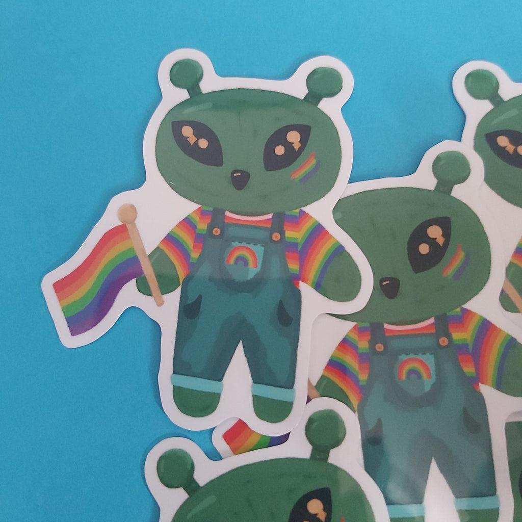 Three cartoon alien stickers with green bodies, rainbow-striped shirts, and blue overalls arranged on a bright blue background.