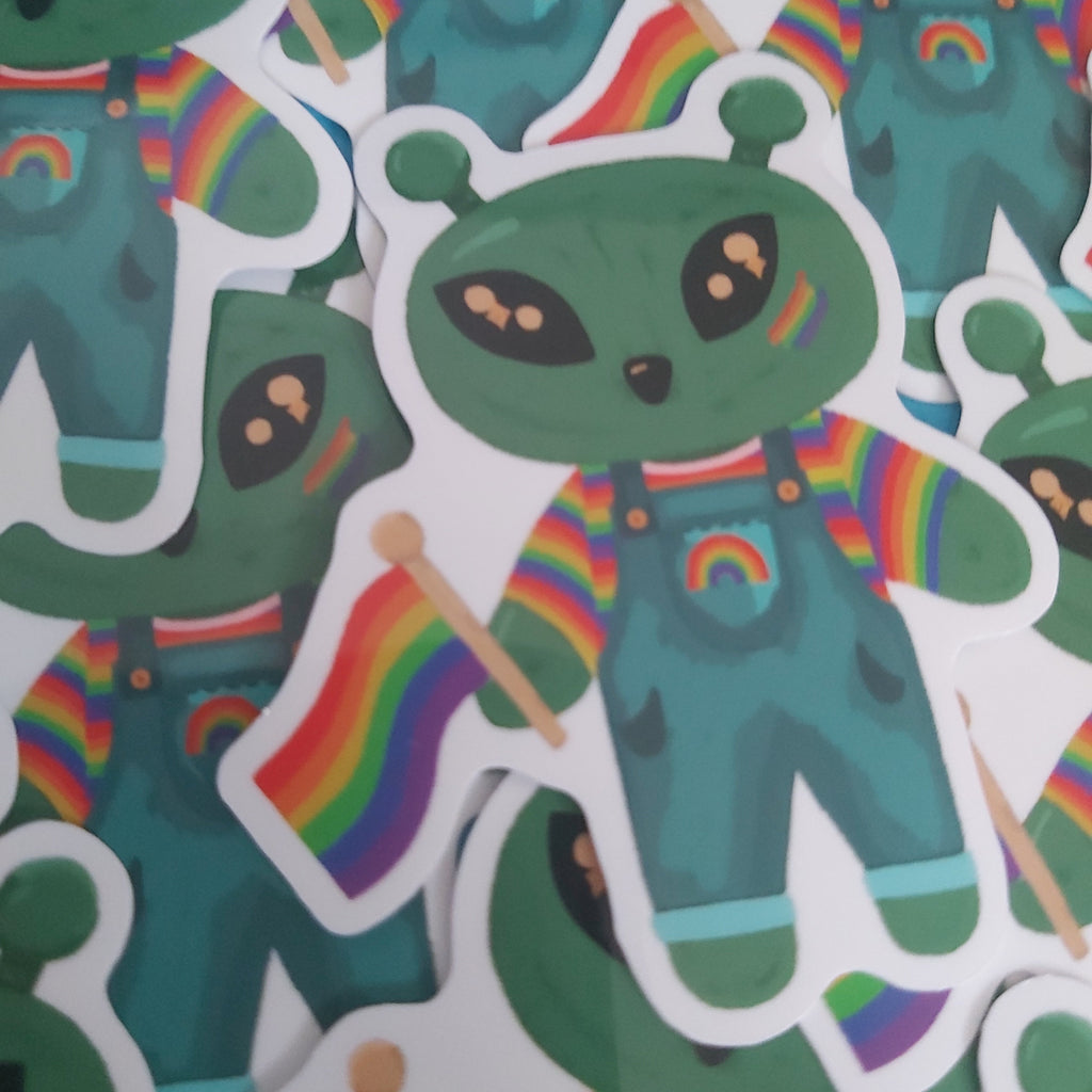 A close-up of multiple die-cut stickers featuring a green alien character wearing teal overalls and holding a rainbow flag, arranged on a white background.