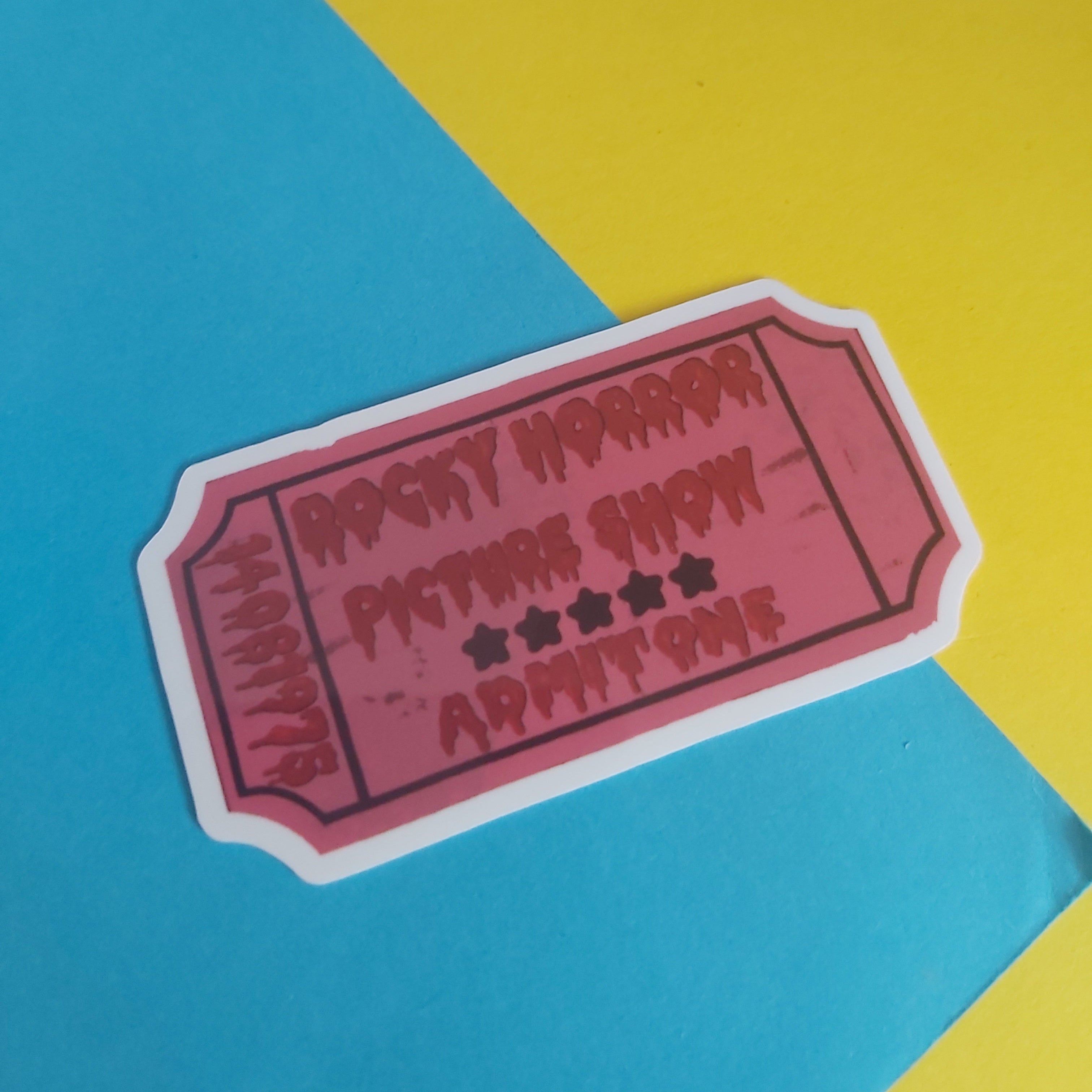 A die-cut sticker shaped like a vintage movie ticket, featuring the text 'ROCKY HORROR PICTURE SHOW' with star ratings, shown on a diagonally split yellow and blue background.