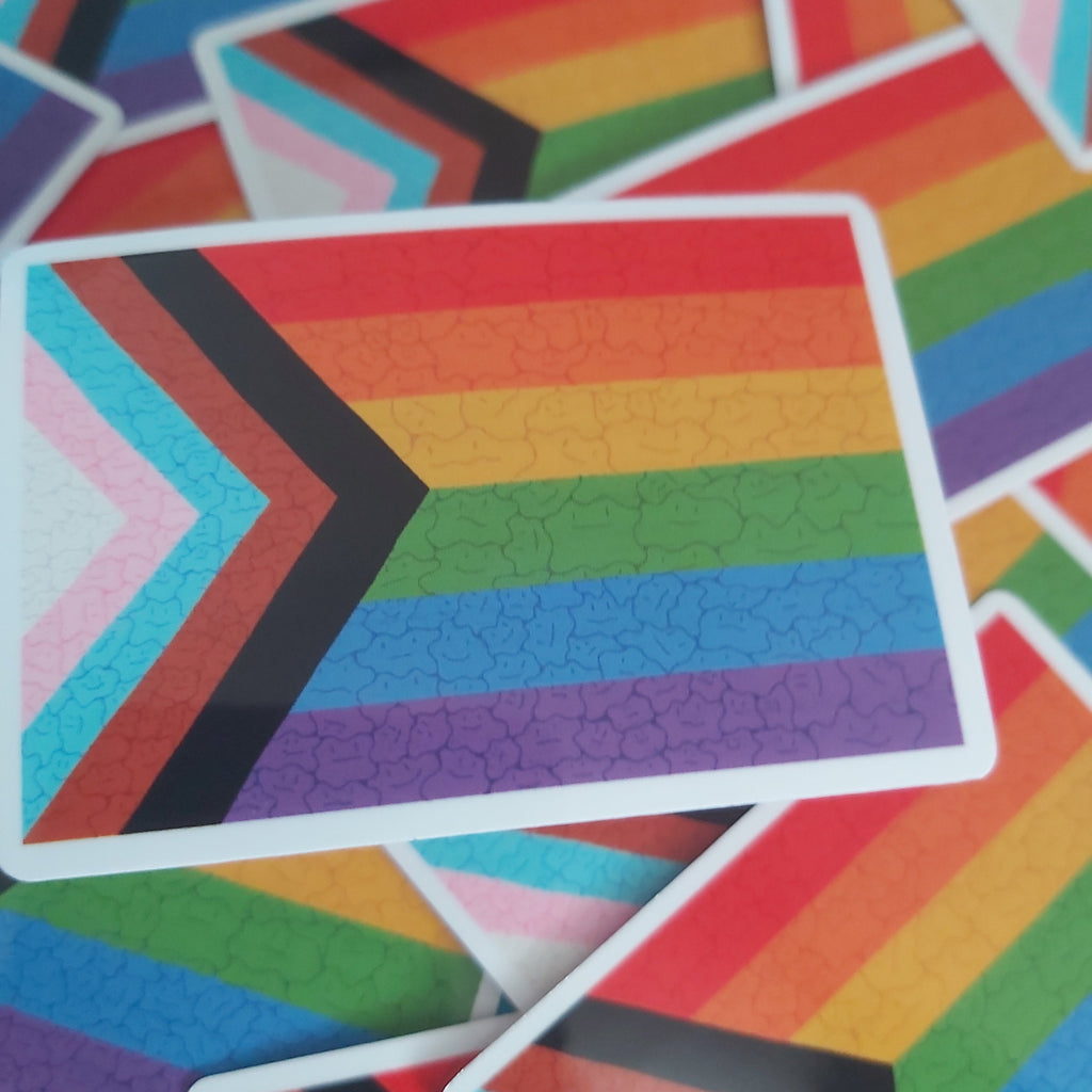 A close-up of a rectangular sticker featuring the Progress Pride flag design with rainbow stripes and chevron accents, surrounded by overlapping similar stickers.