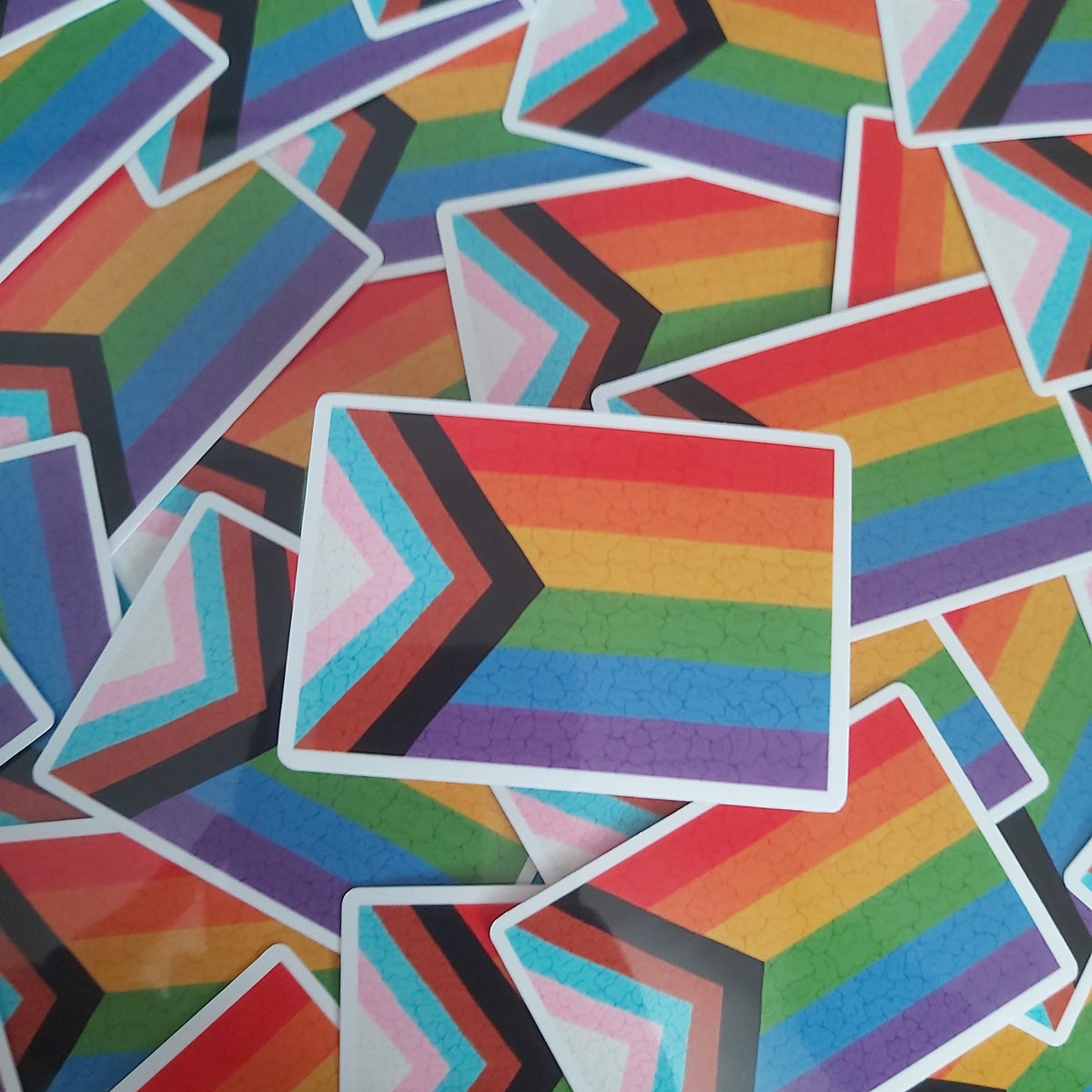 A pile of small rectangular stickers featuring the Progress Pride flag chevron pattern, with rainbow stripes and black, pink, light blue, and white chevron bands.