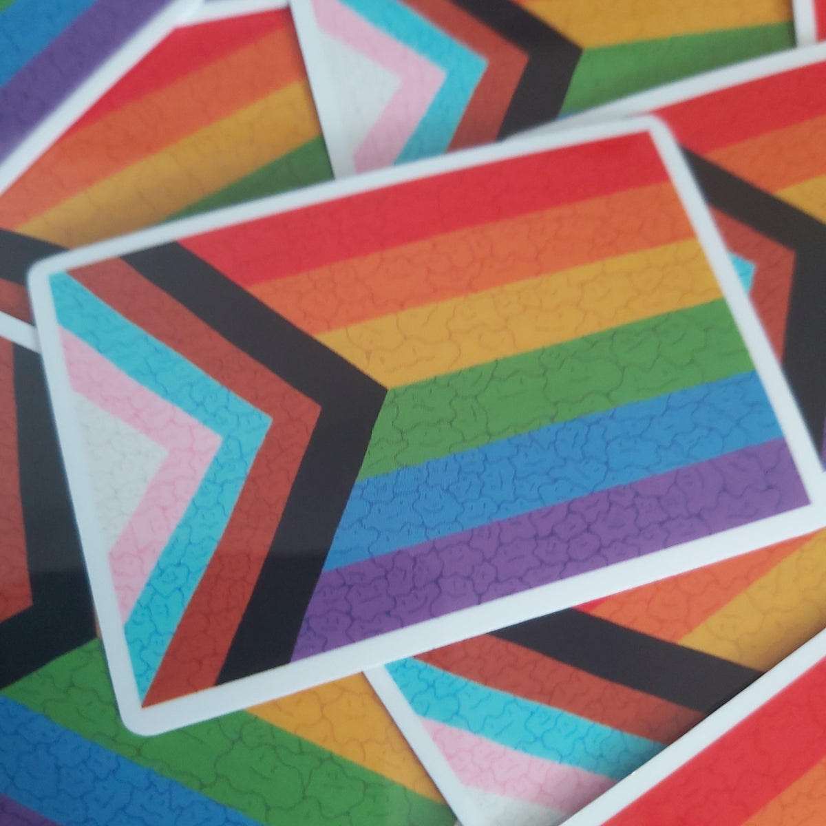 A close-up of several rectangular stickers featuring the Progress Pride flag design, with rainbow stripes and chevron bands in white, pink, light blue, black, orange, yellow, green, blue, and purple.