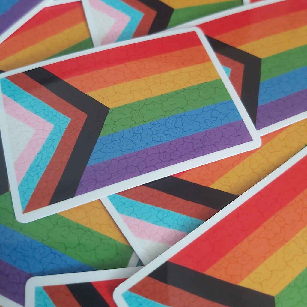 A close-up pile of rectangular stickers featuring the Progress Pride Flag design with subtle ditto faces, showing overlapping rainbow stripes and chevron bands in pink, light blue, turquoise, purple, black, red, orange, yellow, and green.