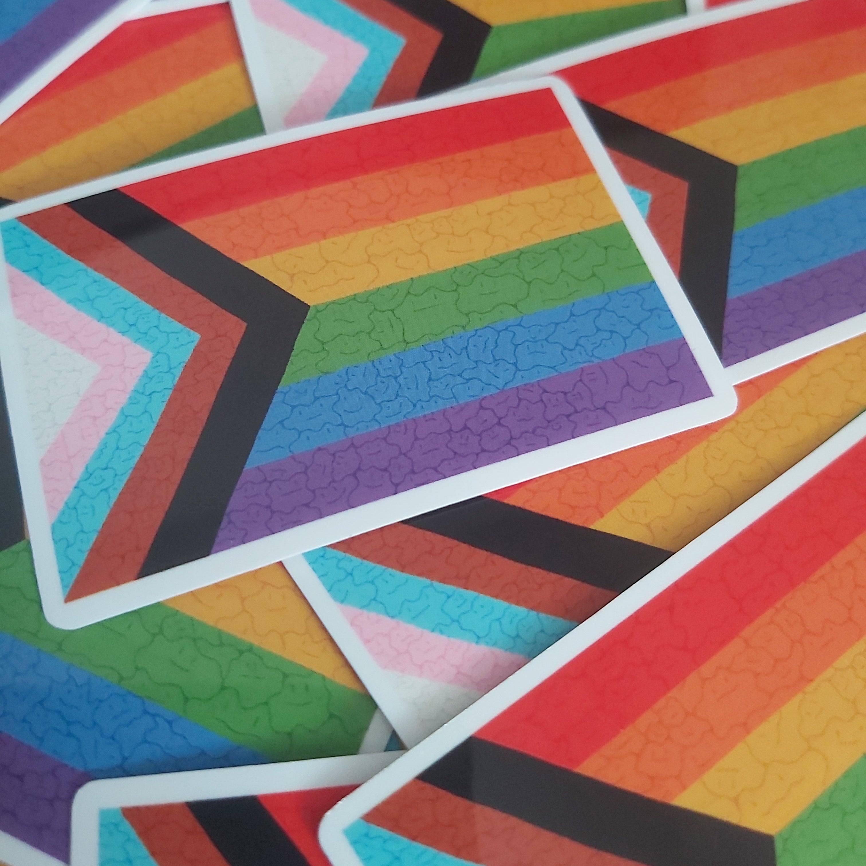 A close-up pile of rectangular stickers featuring the Progress Pride Flag design with subtle ditto faces, showing overlapping rainbow stripes and chevron bands in pink, light blue, turquoise, purple, black, red, orange, yellow, and green.