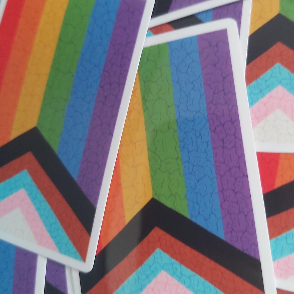 A close-up of several colourful geometric stickers scattered on a surface. The design combines rainbow vertical stripes on the top half with chevron bands in black, red, orange, pink, light blue, and white on the bottom half.