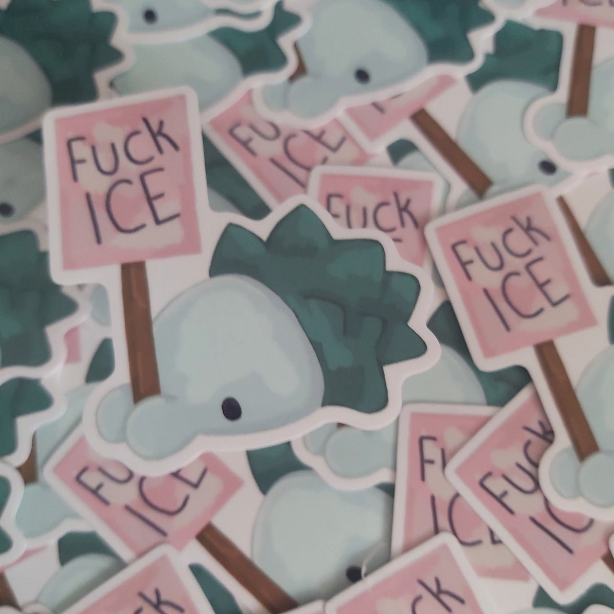 A pile of die-cut stickers featuring a pokemon holding a pink sign that reads 'Fuck ICE' in black handwritten-style text. The stickers are layered together, showing the white borders and other overlapping designs.