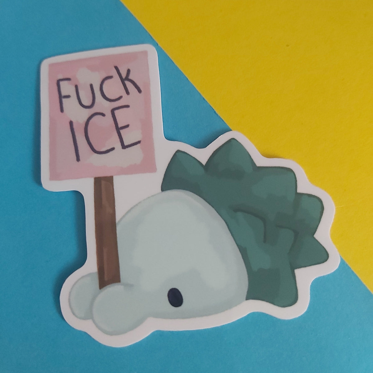 A die-cut sticker featuring a cute creature holding a pink sign that reads 'FUCK ICE' in black handwritten text. The sticker is displayed on a diagonally split blue and yellow background.