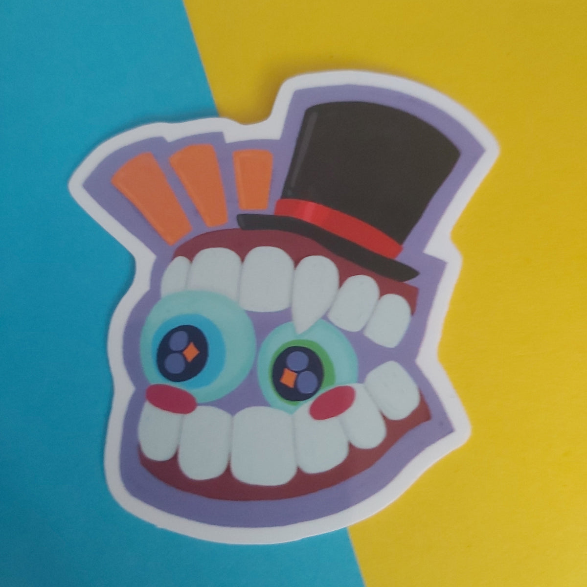 A colourful die-cut sticker featuring a stylised cartoon face with large round eyes, white teeth, a black top hat with a red band on a purple background, shown on a split blue and yellow backdrop.