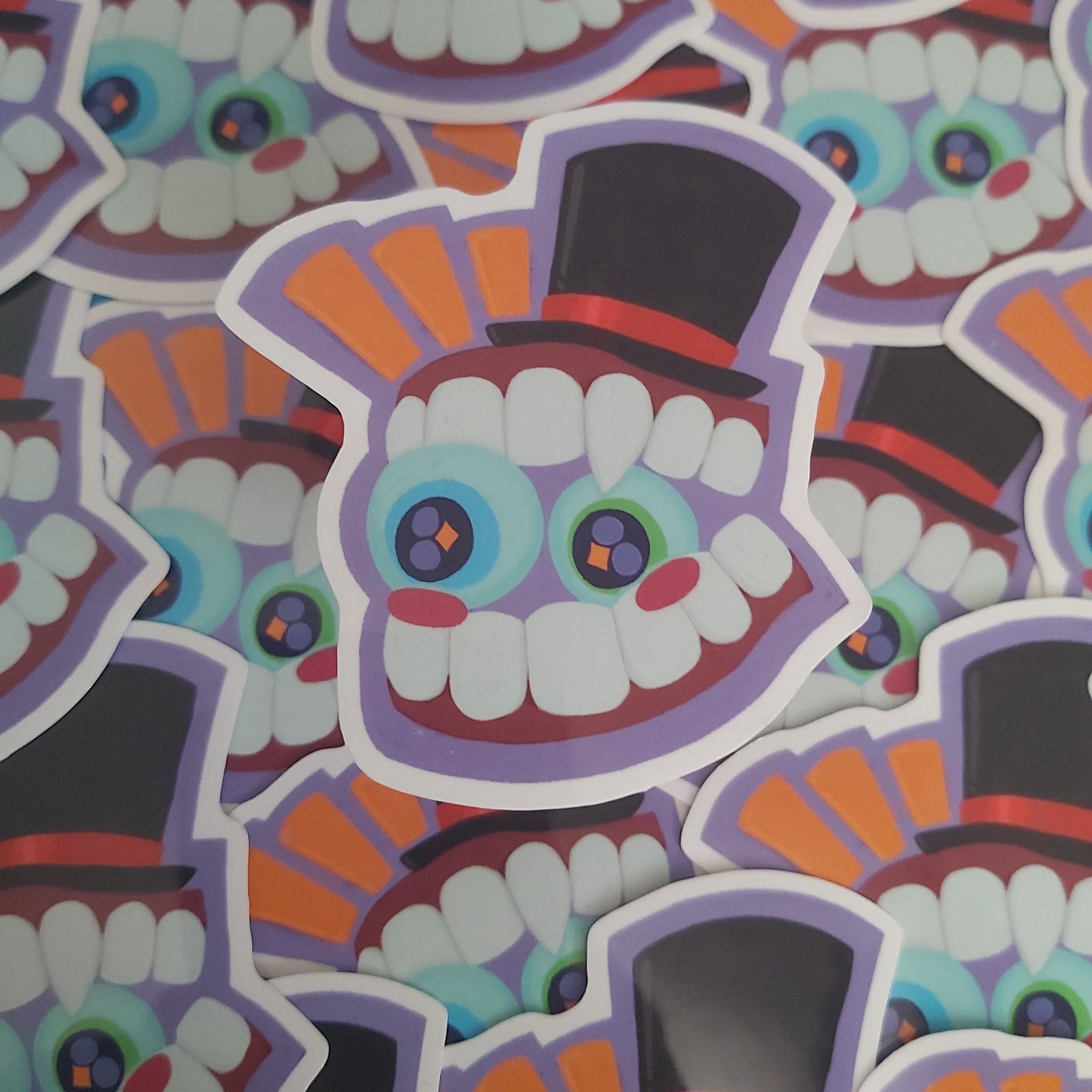 A close-up pile of die-cut stickers featuring a stylised cartoon toothface with large blue and green eyes, white teeth, purple outlines, orange and black top hat details, and red cheeks.