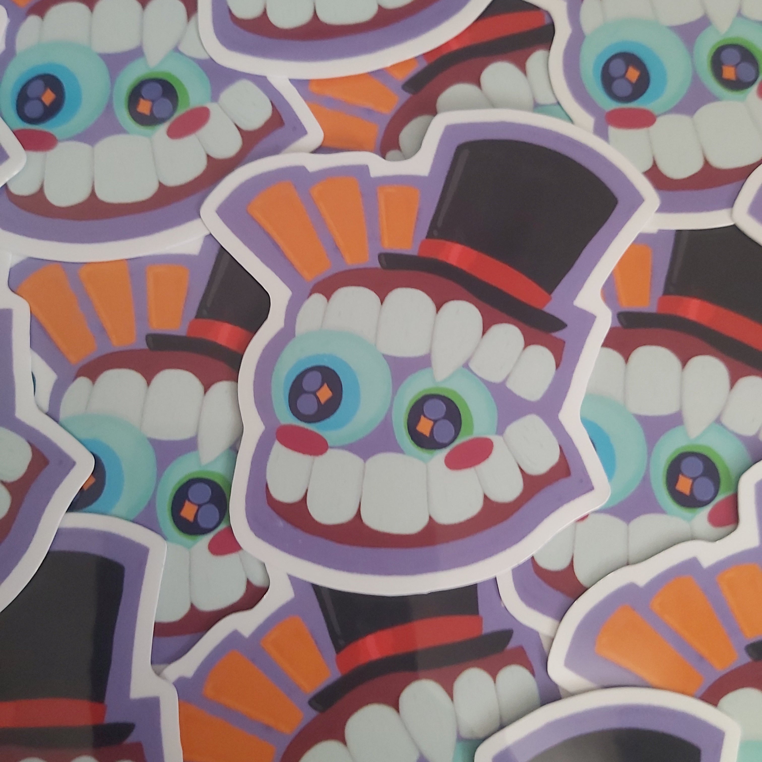 A pile of colorful die-cut stickers featuring a stylized cartoon face with large eyes, white teeth, and a black top hat with orange rays behind it.