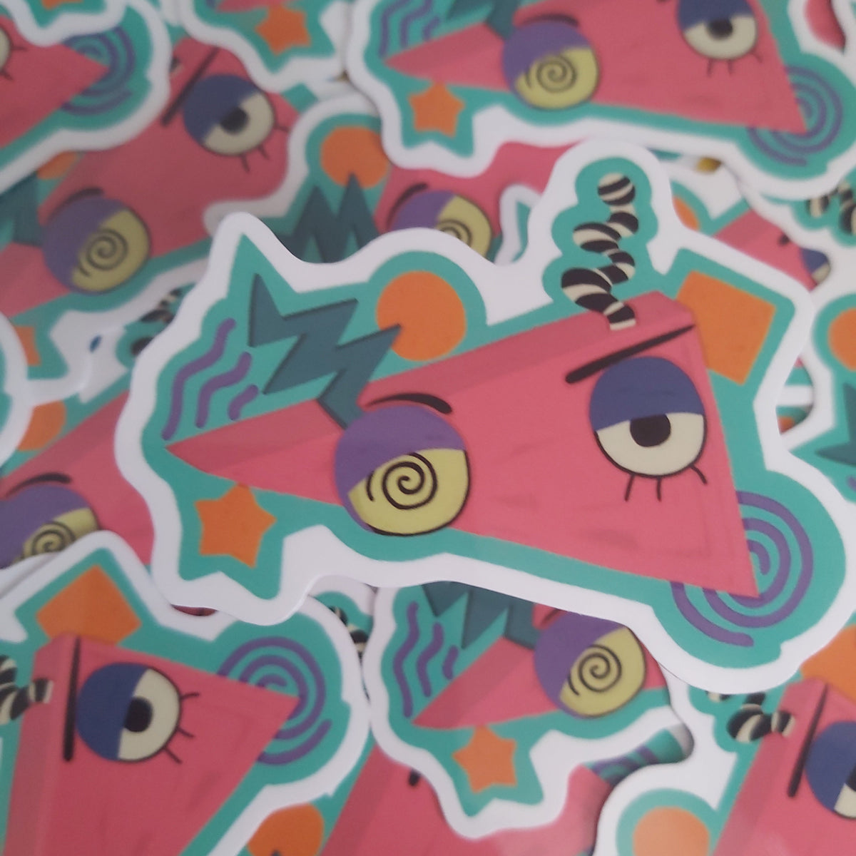 A pile of colourful die-cut stickers featuring a pink triangular cartoon face with teal and yellow accents.