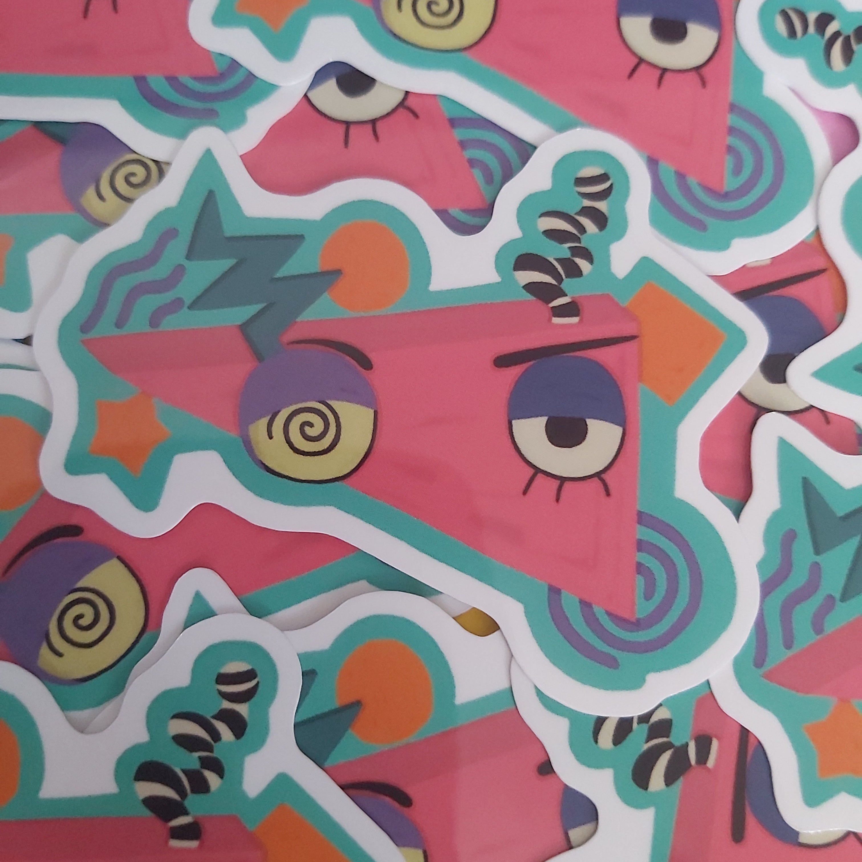 A pile of colourful die-cut stickers featuring a whimsical cartoon with a pink triangle face, large expressive eyes, and abstract shapes in teal, purple, and yellow.
