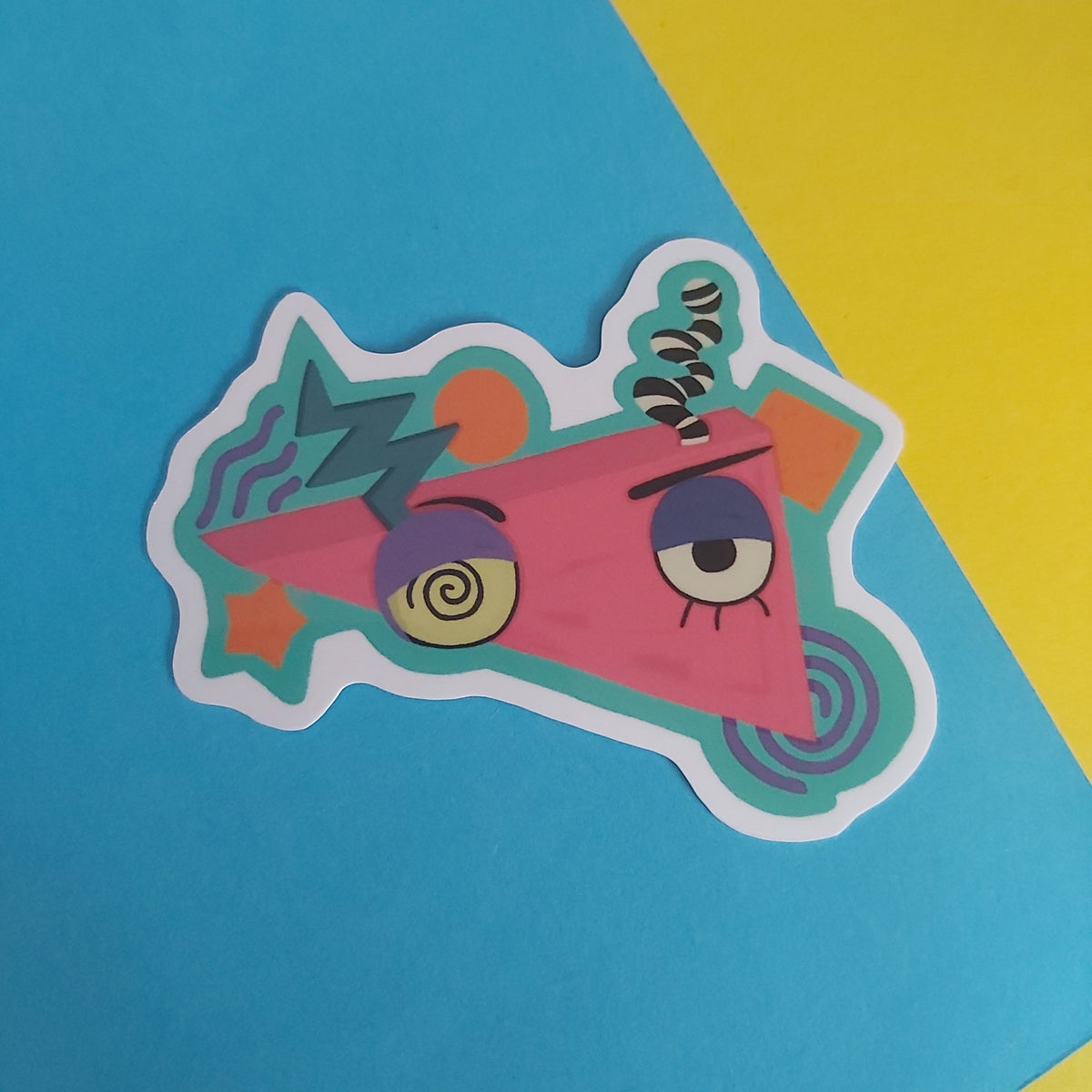 A colorful die-cut sticker featuring a cartoon triangle character with a spiral eye and a coiled spring, placed on a diagonally split blue and yellow background.