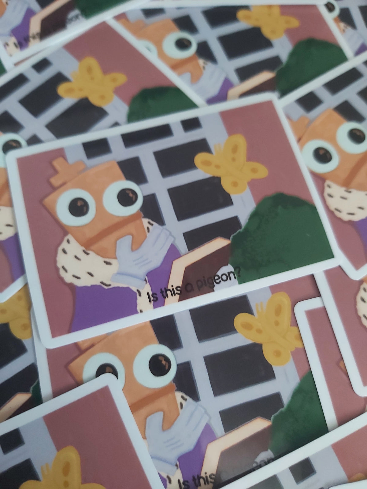 A close-up of several printed stickers scattered on top of each other. The artwork shows a cartoon chess piece with large eyes, a purple shirt, a green bush, and a yellow butterfly.