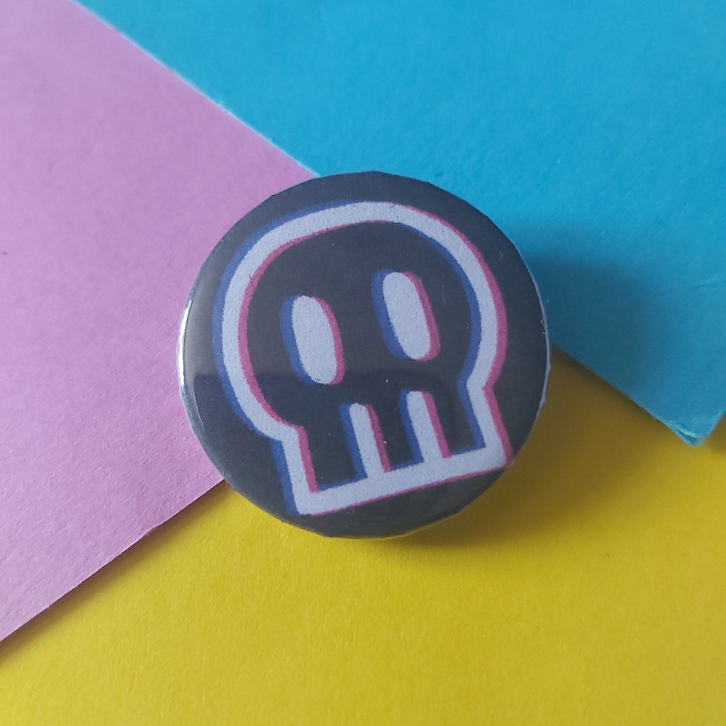 A round pin badge featuring a stylized white skull outline with pink and blue glitch-style borders on a dark gray background, resting on a diagonally split yellow, pink, and blue paper surface.