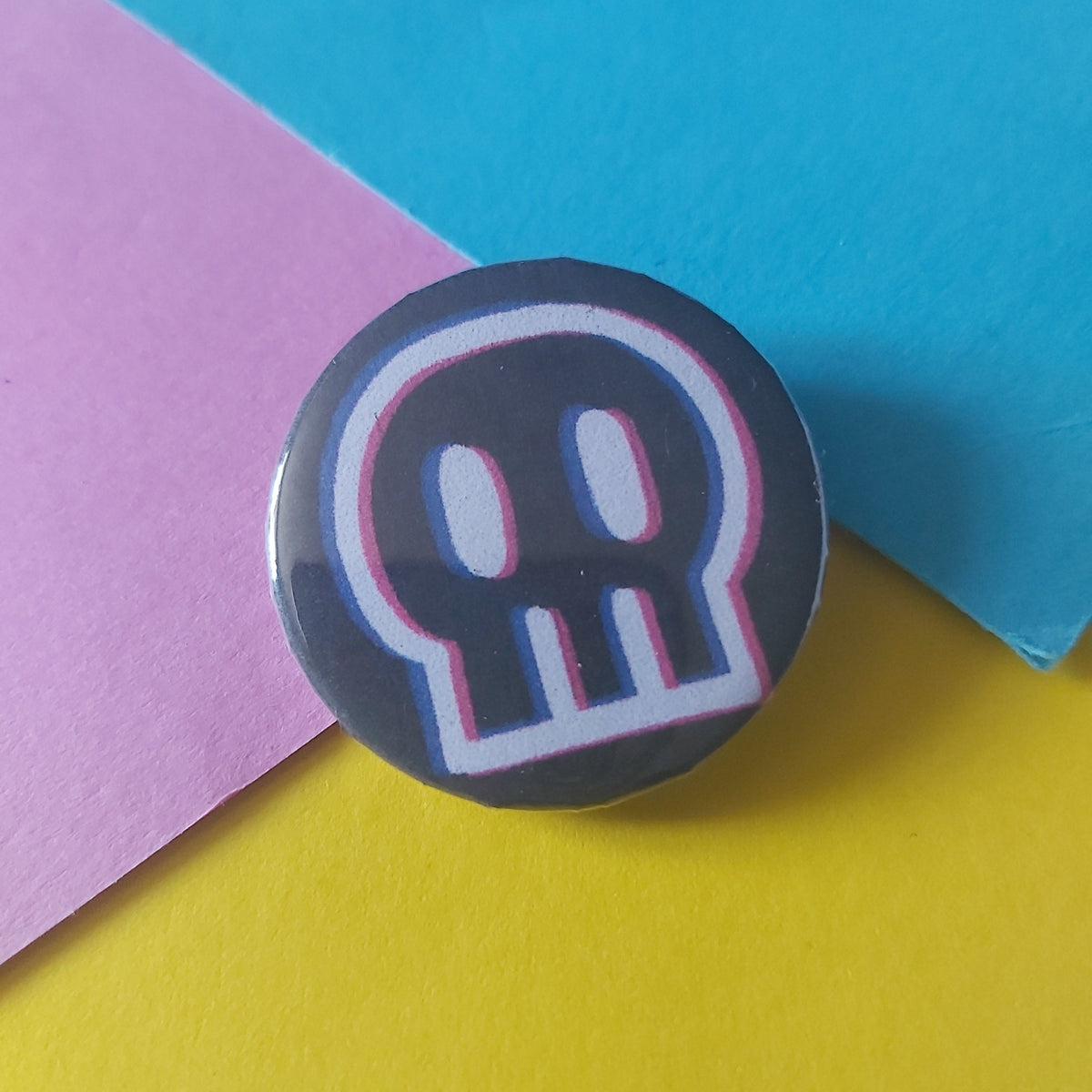 A round pin badge featuring a stylized white skull outline with pink and blue glitch-style borders on a dark gray background, resting on a diagonally split yellow, pink, and blue paper surface.
