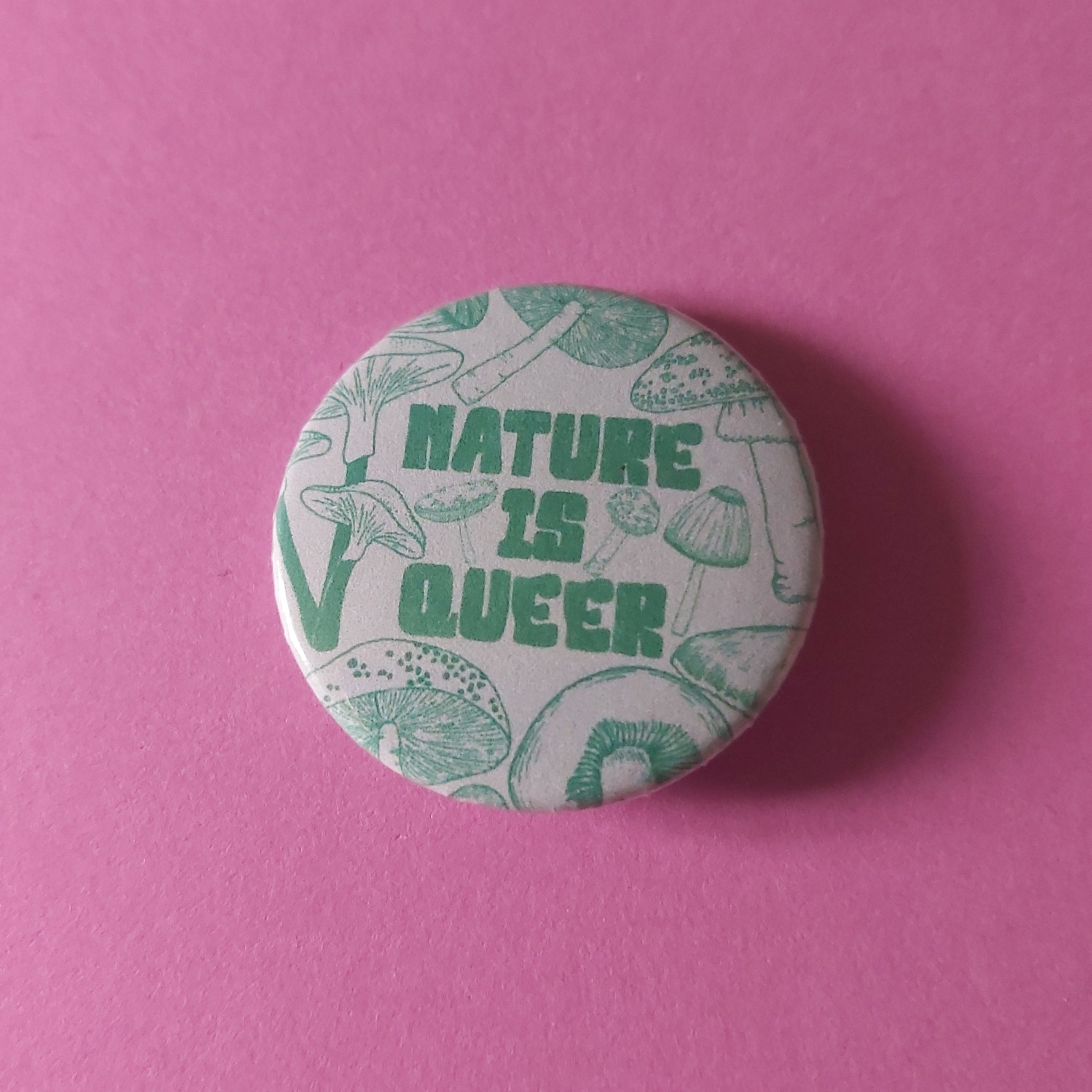 A round pinback button badge featuring the text 'NATURE IS QUEER' in green, surrounded by mushroom illustrations on a white background, set against a solid pink backdrop.