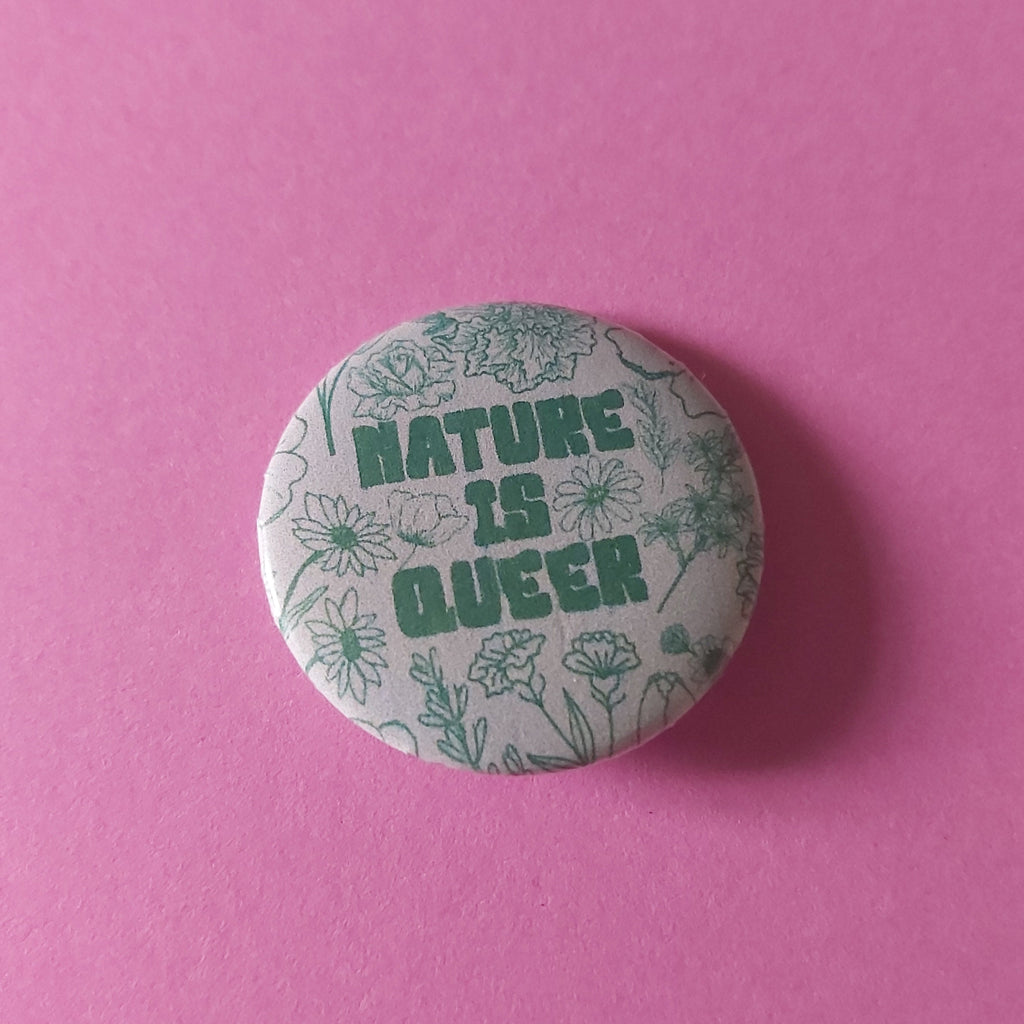 A round pinback button badge featuring the text 'NATURE IS QUEER' in green, surrounded by a light green floral line-art pattern on a white background, set against a solid pink backdrop.