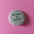 A round pinback button badge featuring the text 'NATURE IS QUEER' in green, surrounded by a light green floral line-art pattern on a white background, set against a solid pink backdrop.