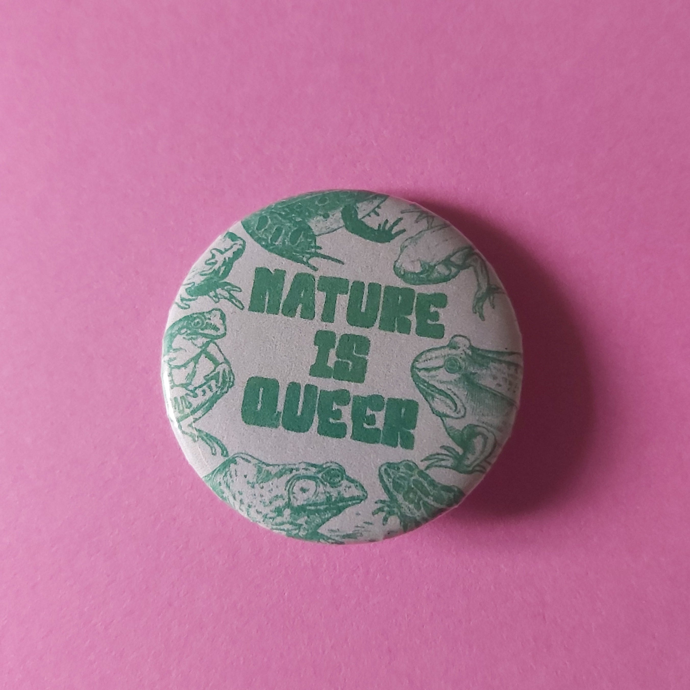 A round pinback button featuring the text 'NATURE IS QUEER' in green, surrounded by green frog illustrations on a white background, set against a solid pink backdrop.