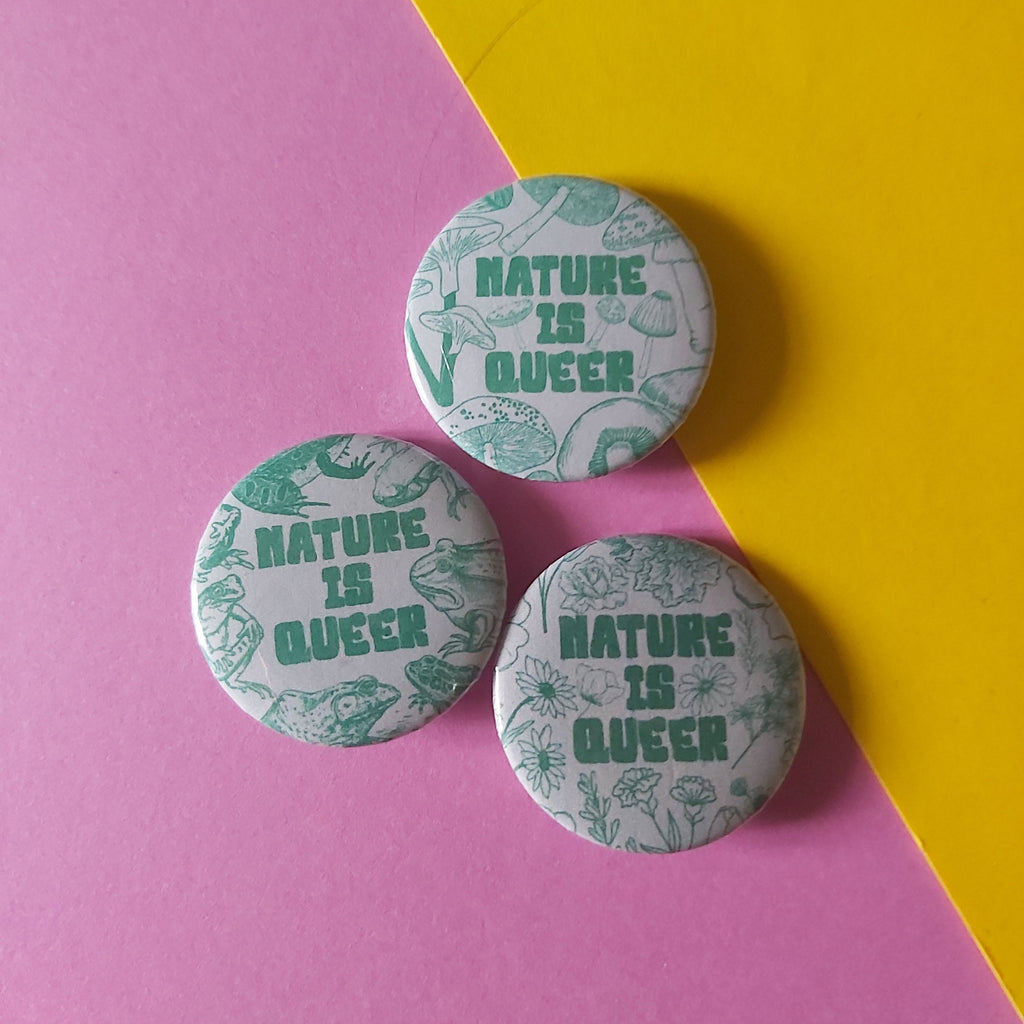 Three round pin-back buttons with the phrase “NATURE IS QUEER” in green text over a pale patterned background, arranged on a diagonally split pink and yellow surface.