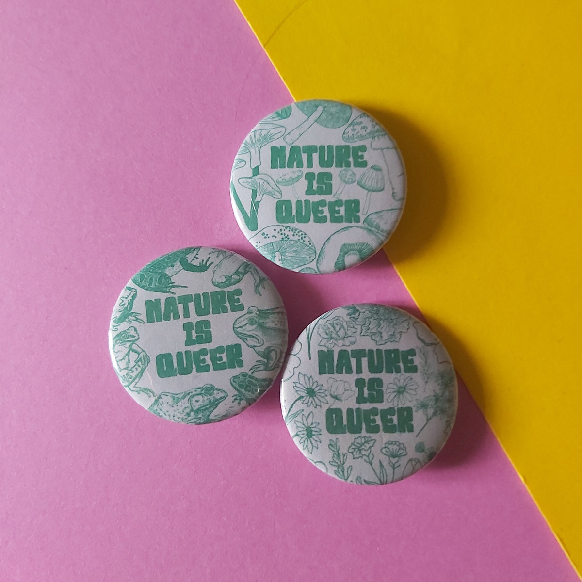 Three round pin-back buttons with the phrase “NATURE IS QUEER” in green text over a pale patterned background, arranged on a diagonally split pink and yellow surface.
