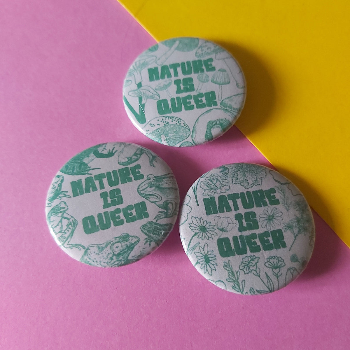 Three round pin-back buttons with a green botanical and frog pattern on a white background, featuring the text 'NATURE IS QUEER' in green, arranged on a diagonally split pink and yellow background.