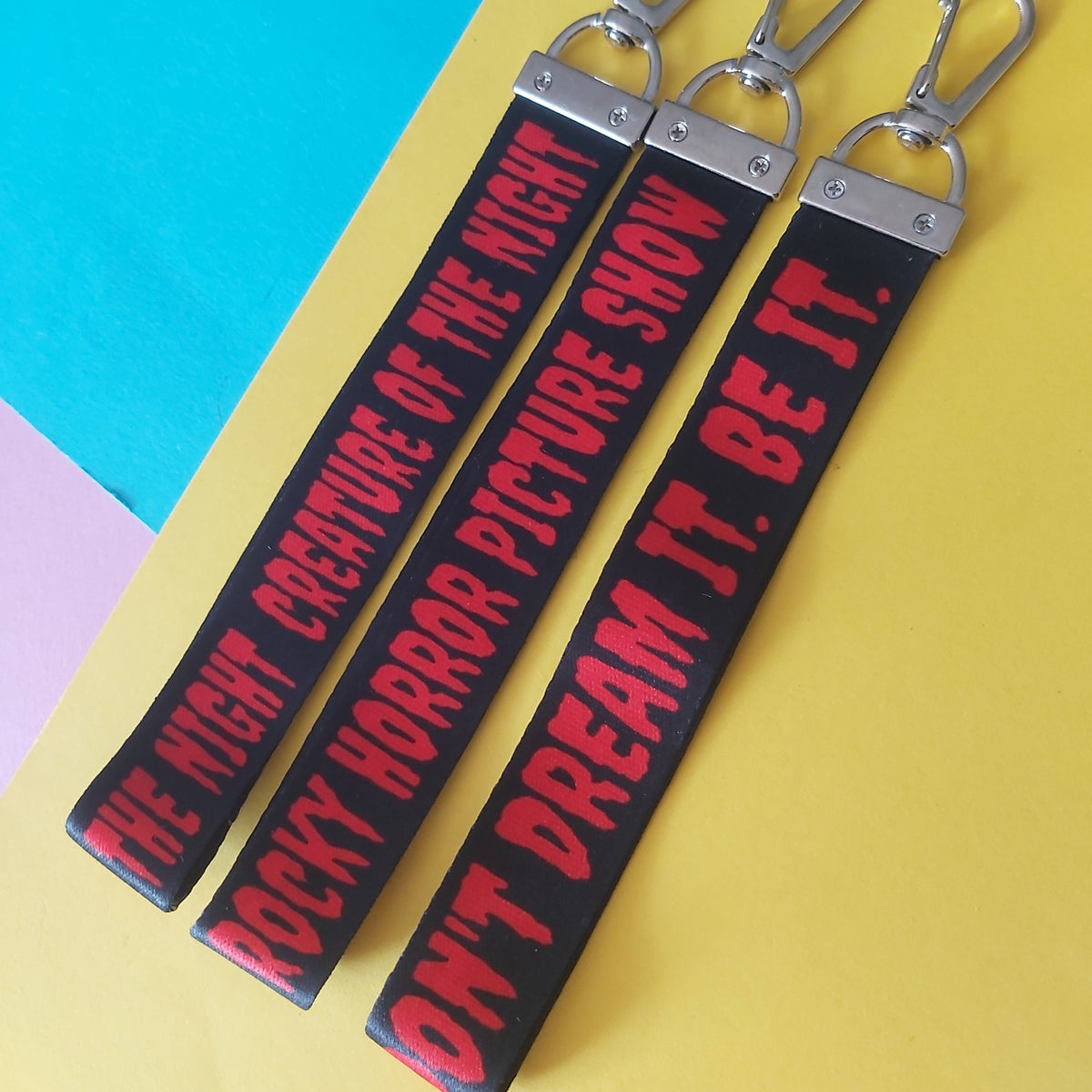 Three black fabric keychain straps with silver metal clasp hardware, printed with red horror-themed text, arranged diagonally on a yellow and blue background.