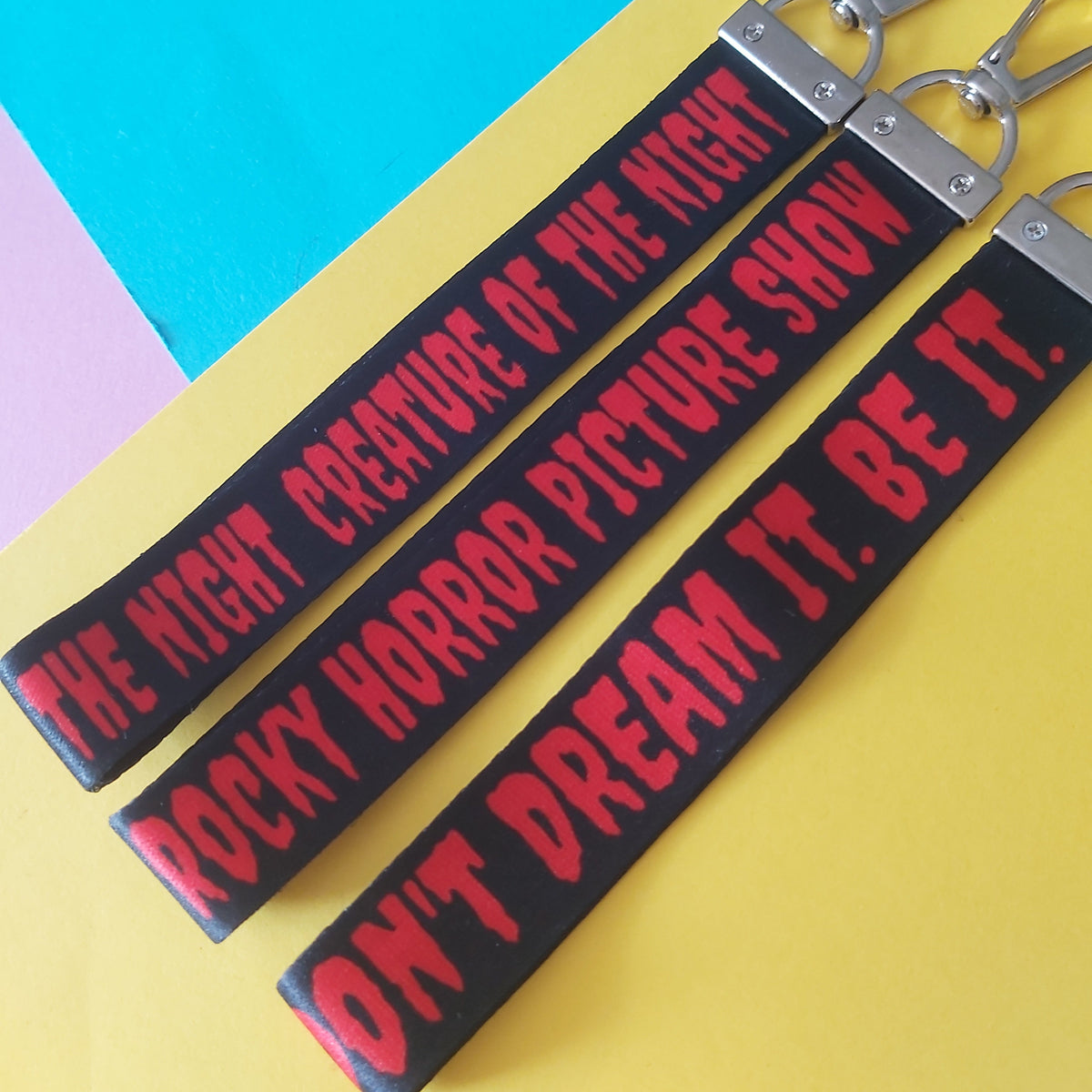 Three black fabric keychains with silver metal lobster clasps, printed with red horror-style text reading 'CREATURE OF THE NIGHT', 'ROCKY HORROR PICTURE SHOW', and 'DON'T DREAM IT. BE IT.