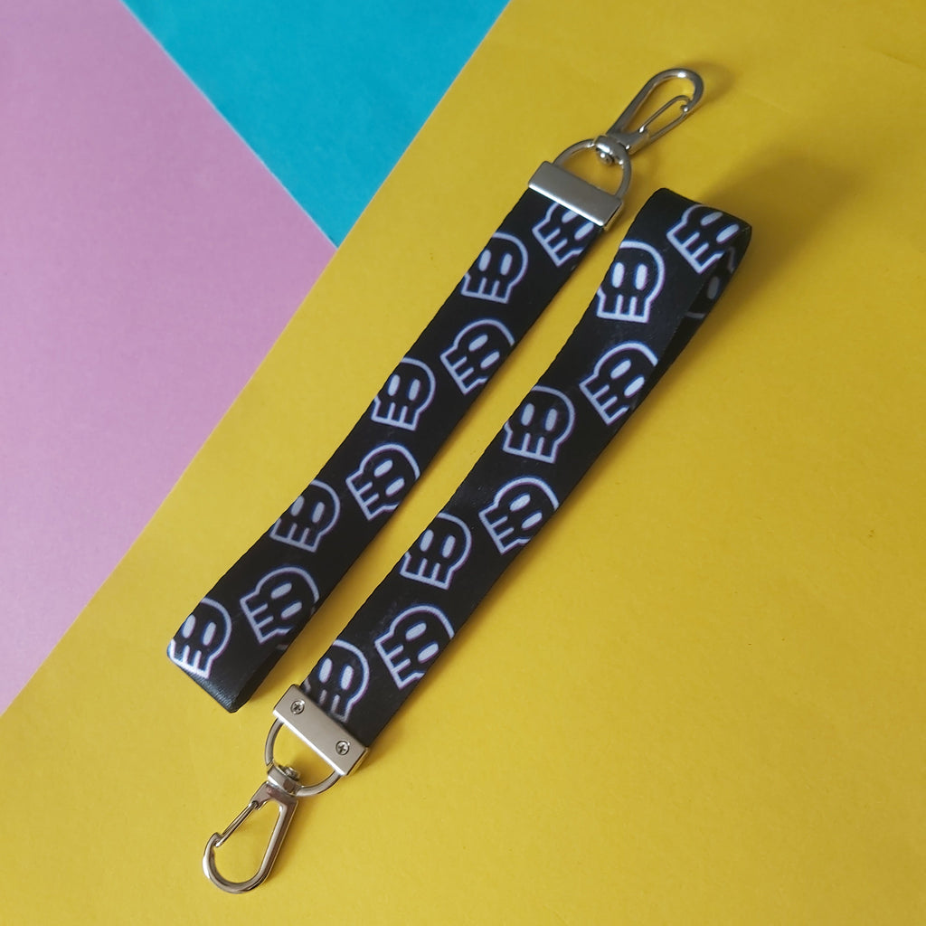 A black fabric wristlet keychain strap with a repeating white skull pattern and silver metal clasps, laid diagonally on a bright yellow background with pink and teal paper sections visible in the corners.