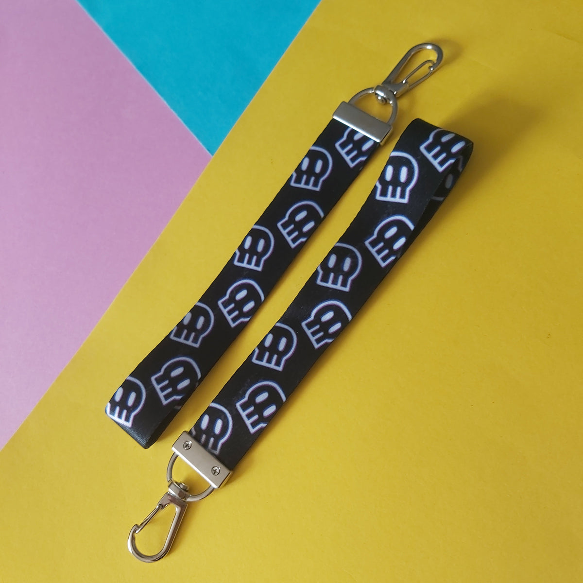 A black fabric wristlet keychain strap with a repeating white skull pattern and silver metal clasps, laid diagonally on a bright yellow background with pink and teal paper sections visible in the corners.