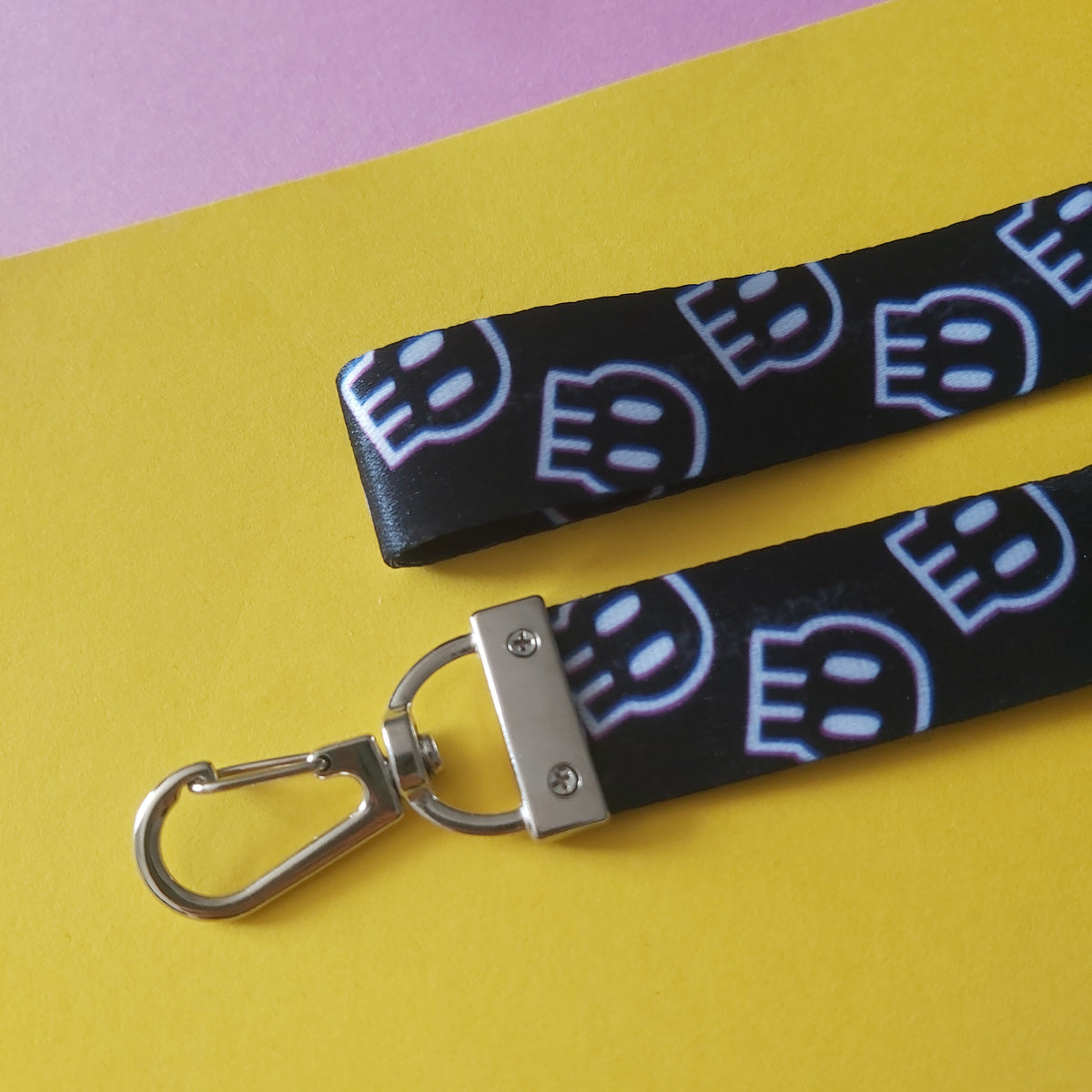A close-up of a black fabric wrist lanyard with a repeating white outlined 'skull pattern and a silver metal swivel clasp, laid on a bright yellow surface with a pink corner visible.