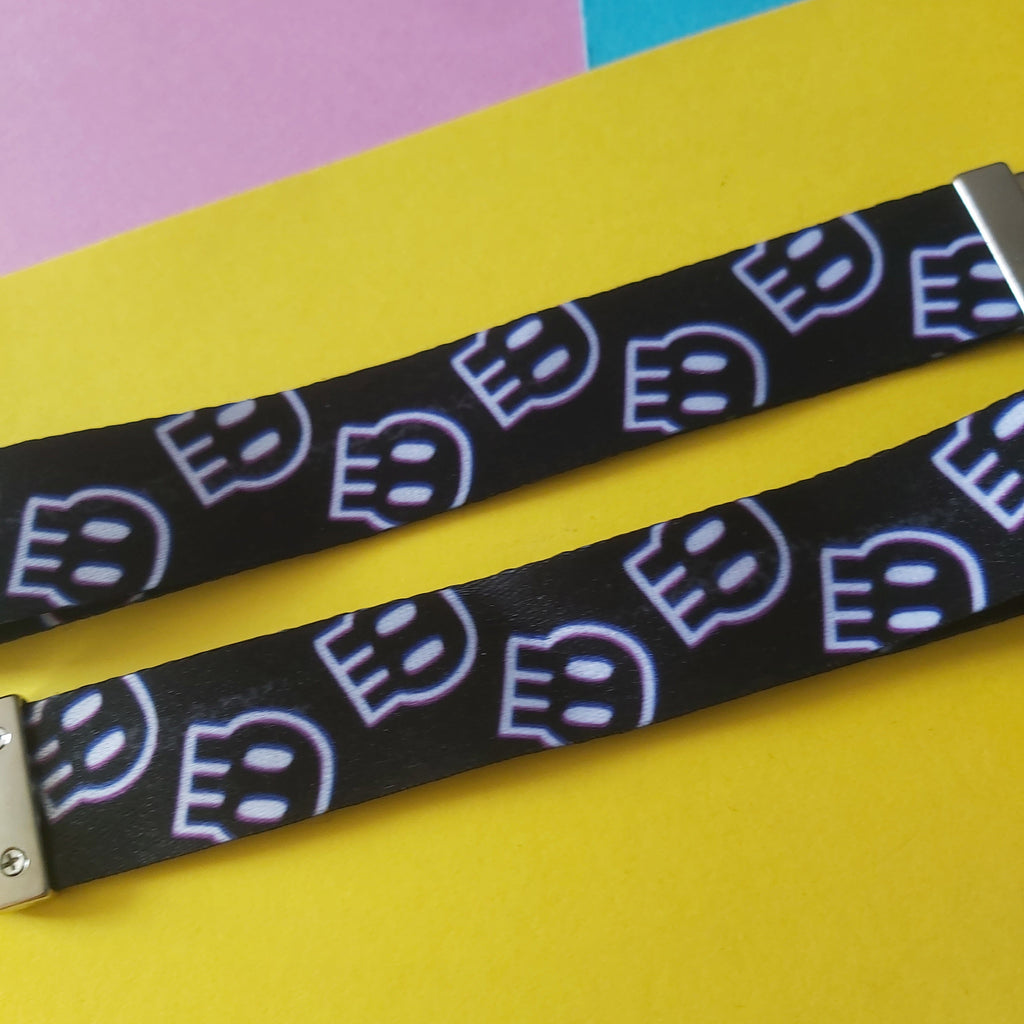 A close-up of a black lanyard strap featuring a repeating pattern of stylised white and light blue skulls, laid diagonally on a bright yellow background with pink and blue paper sections in the upper corner.