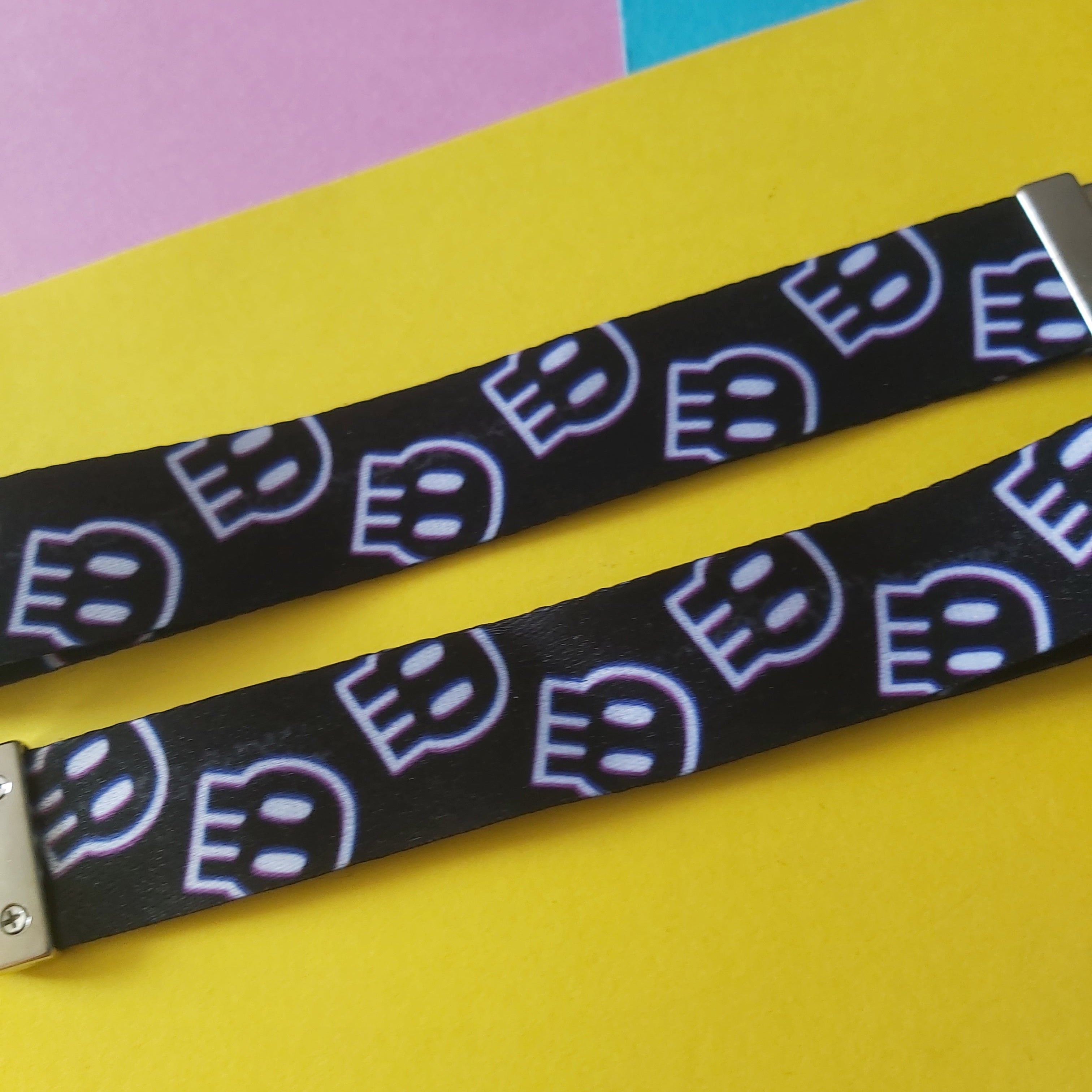 A close-up of a black lanyard strap featuring a repeating pattern of stylised white and light blue skulls, laid diagonally on a bright yellow background with pink and blue paper sections in the upper corner.