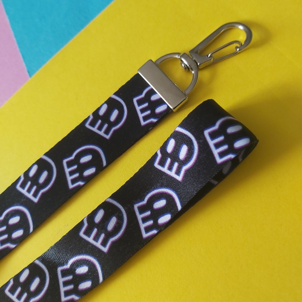 A black fabric lanyard strap with a silver metal swivel clasp, featuring a repeating white and purple skull pattern, displayed on a bright yellow background with a colourful pastel pink and blue corner accent.
