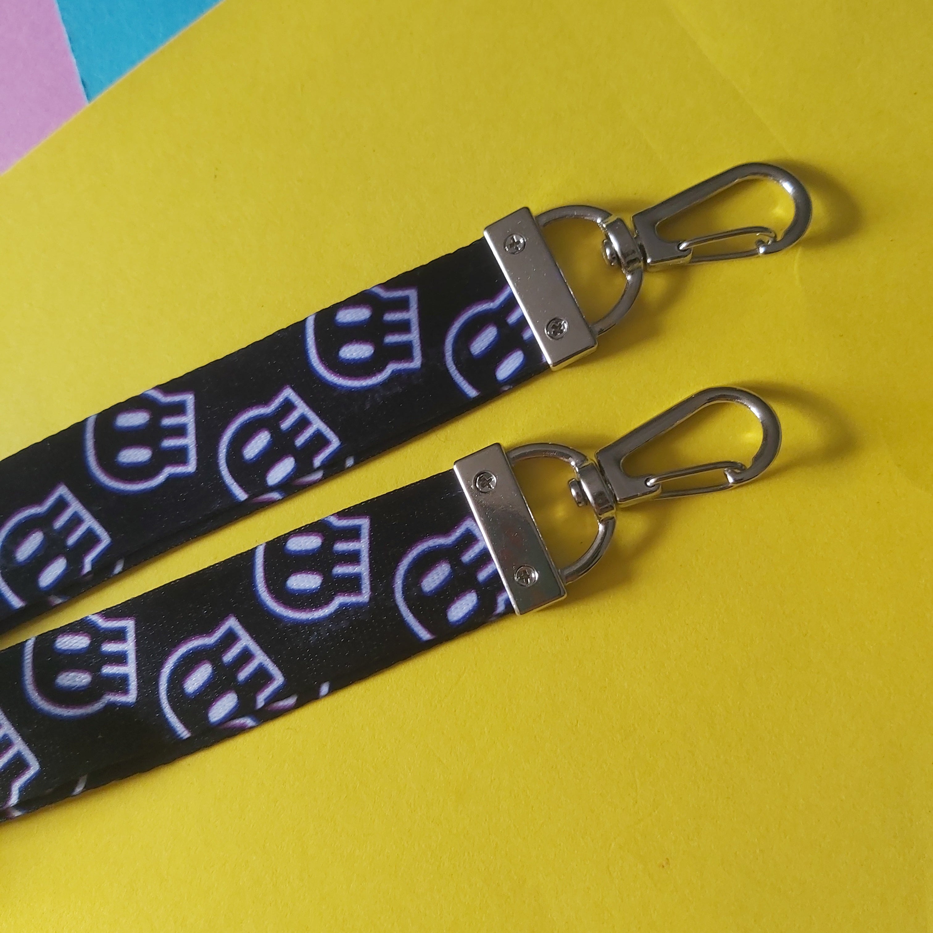 Two black fabric lanyards with a repeating purple skull pattern and silver metal swivel clasp, laid diagonally on a bright yellow background.