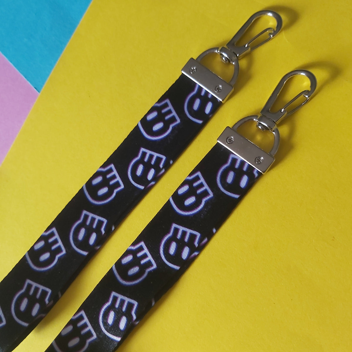 Two black fabric keychains with a repeating white and purple skull pattern, finished with silver metal lobster clasps, displayed diagonally on a bright yellow background with colourful paper corners.