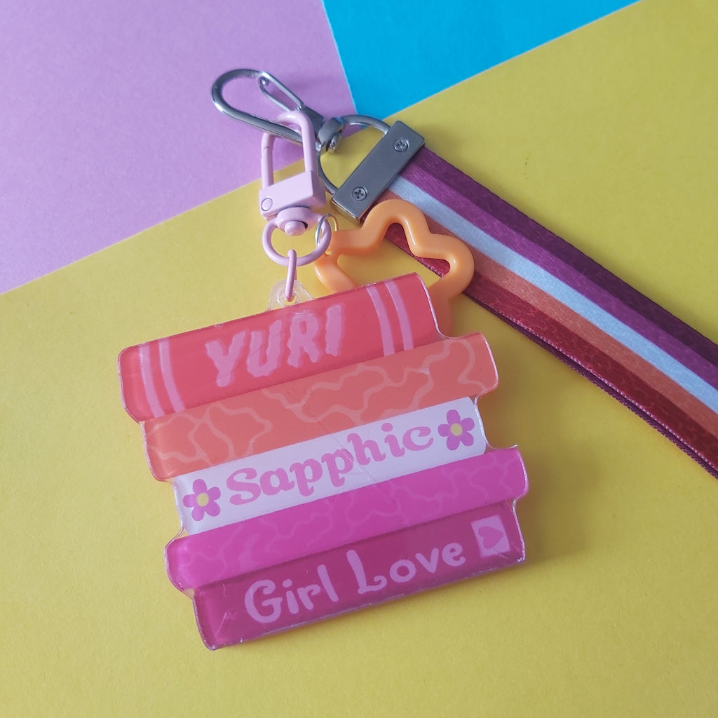 A colourful keychain featuring five stacked book charms with the words 'YURI', 'Sapphic', and 'Girl Love' in white script, attached to a silver clasp and a striped wrist strap on a pastel-colored background.
