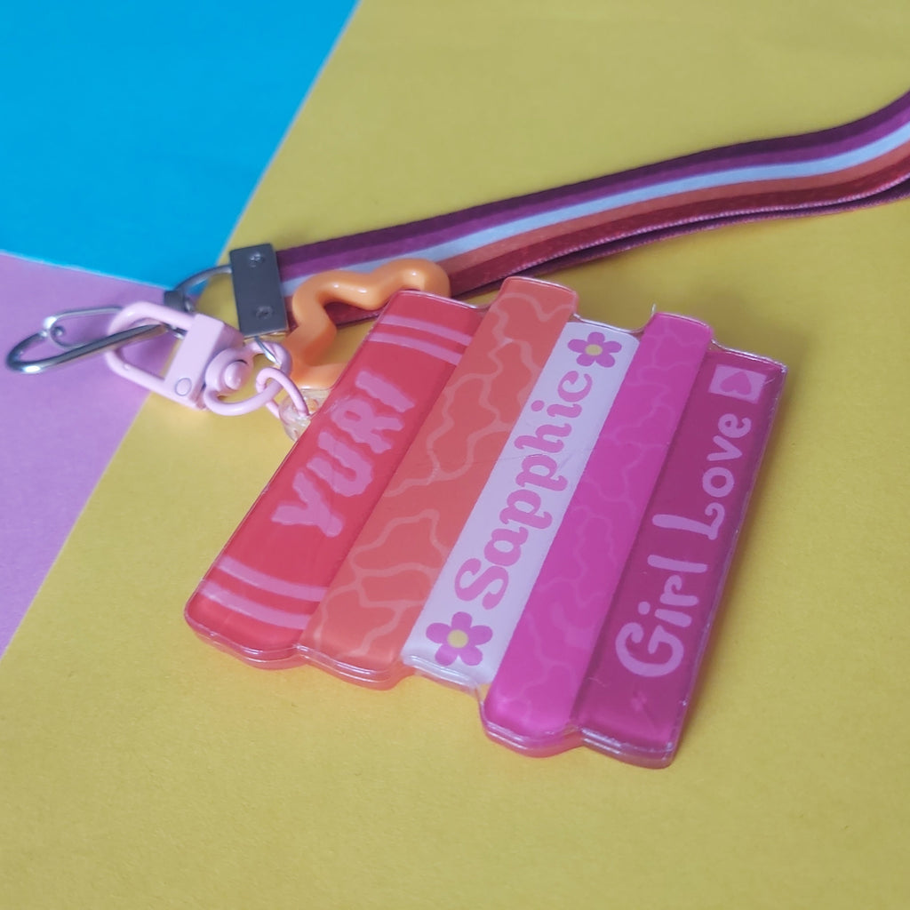 A close-up of a keychain wristlet charm set featuring five colourful, stacked books with the words 'YURI', 'Sapphic', and 'Girl Love' on a striped pink and orange lesbian flag strap. The product is photographed on a background split diagonally between yellow and pastel blue/pink.