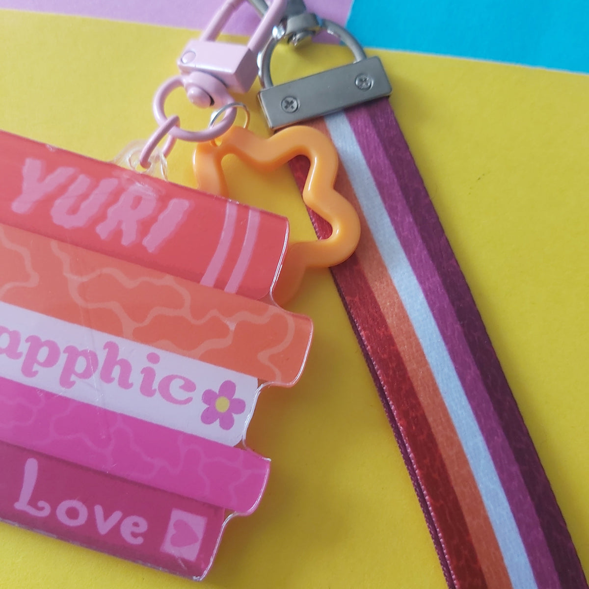 A close-up of a colourful keychain wristlet strap accessory with a lobster clasp, metal ring, orange star charm, and striped fabric strap on a bright yellow, pink, and blue background.