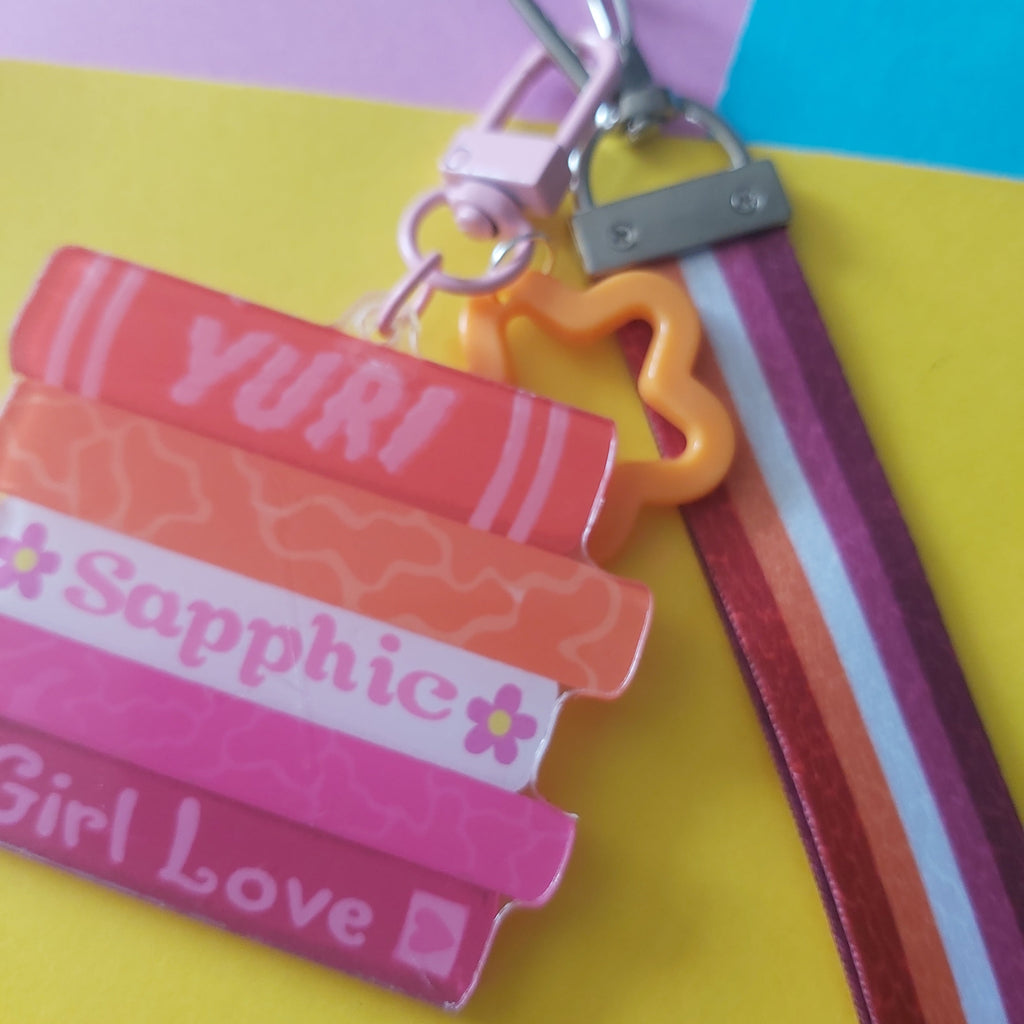 A close-up of a colourful keychain wristlet charm with five stacked book-shaped pieces labelled 'YURI', 'Sapphic', and 'Girl Love', attached to a striped lesbian lanyard and metal clasp on a bright pastel background.