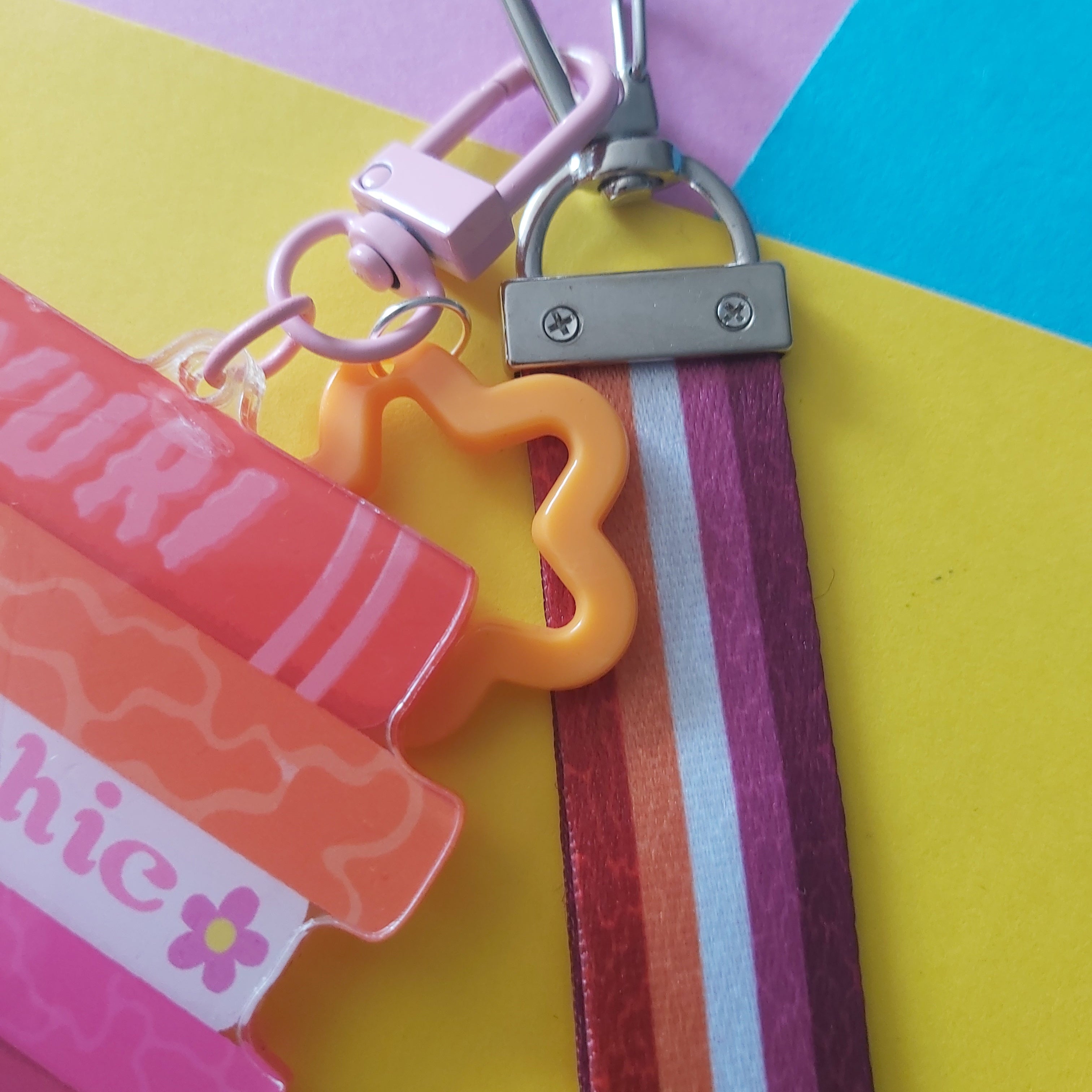 A close-up of a colourful keychain wristlet strap accessory with a silver carabiner, metal connector, striped fabric lesbian strap, and bright plastic charms on a pastel geometric background.