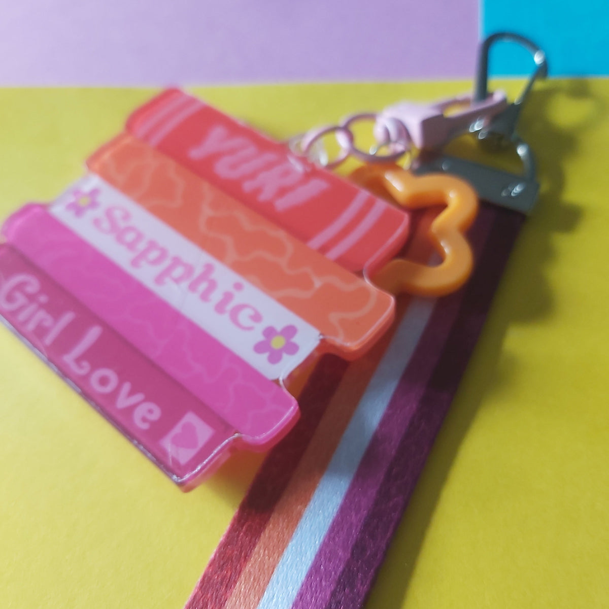 A close-up of a colourful keychain bag charm with a book stack reading 'Yuri', 'Sapphic', and 'Girl Love', attached to a striped pink, orange and white strap with a metal clasp, set against a bright yellow and pastel background.