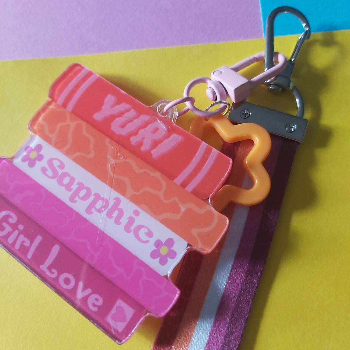 A pastel acrylic keychain bag charm set featuring five stacked book-shaped charms labelled 'YURI', 'Sapphic', and 'Girl Love' in pink, orange, and white, attached to an orange star-shaped charm and a lesbian pride flag-striped strap on a yellow, purple, and blue background.
