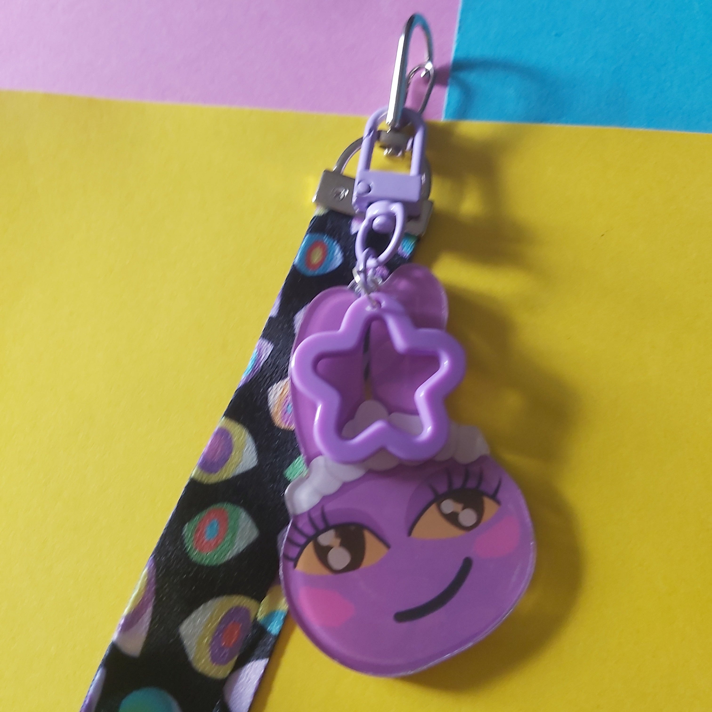 A purple cartoon Jax bunny charm keychain with a star-shaped ring and a black patterned strap, displayed on a yellow and pink background.