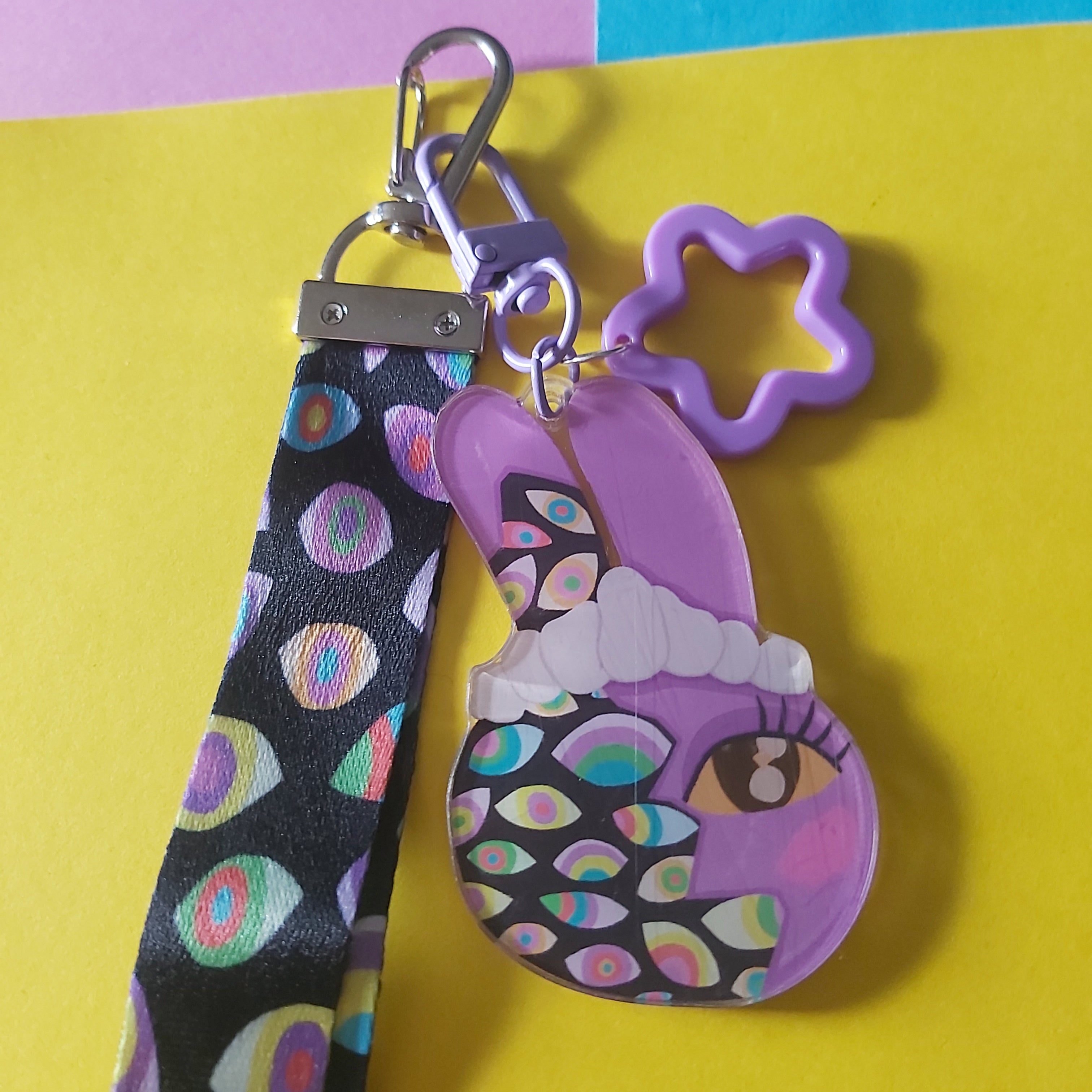 A colourful keychain and wristlet strap set featuring a purple cartoon character charm, a purple star-shaped clip, and a black patterned fabric strap with multicoloured eye motifs, displayed on a yellow and pastel-colored background.