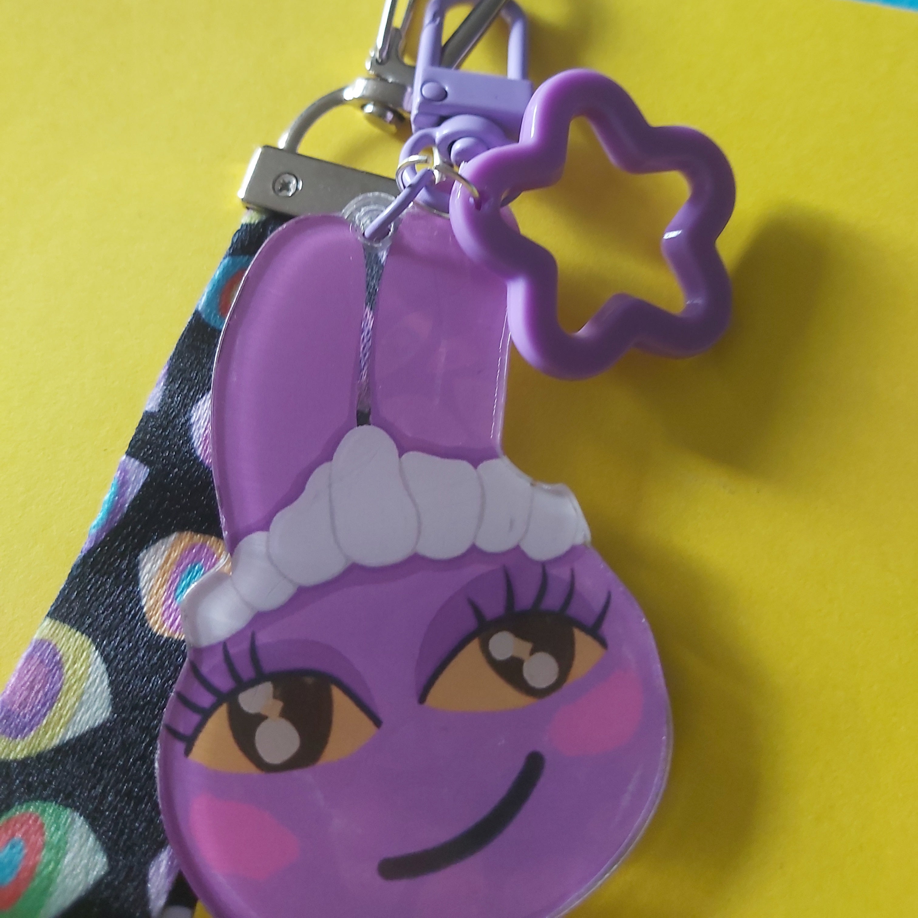 A close-up of a purple cartoon bunny keychain with a metal clasp, a purple star-shaped ring, and a patterned fabric loop, set against a bright yellow and pink background.
