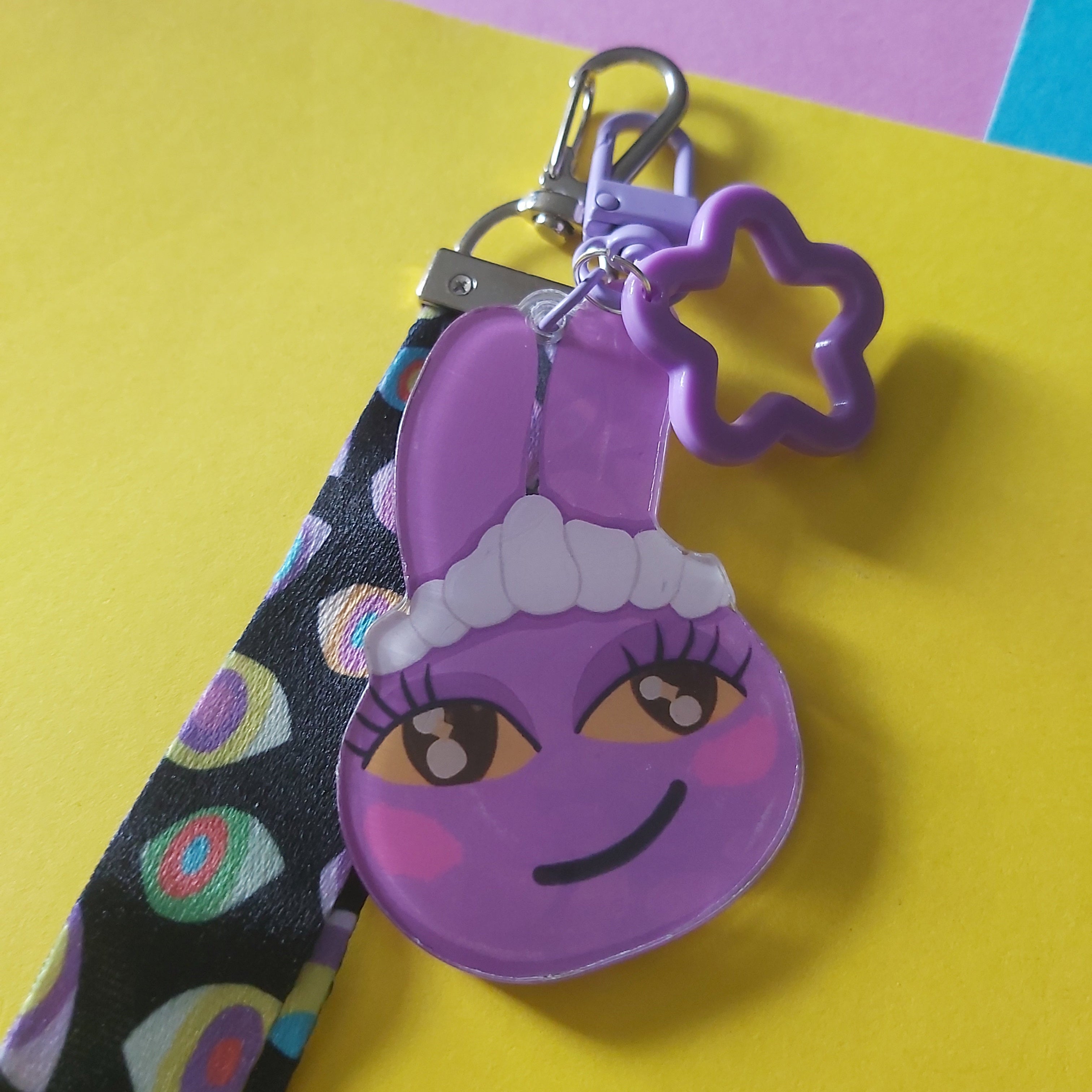 A purple cartoon bunny keychain with a matching wrist strap, resting on a bright yellow paper background with a pink and blue corner.