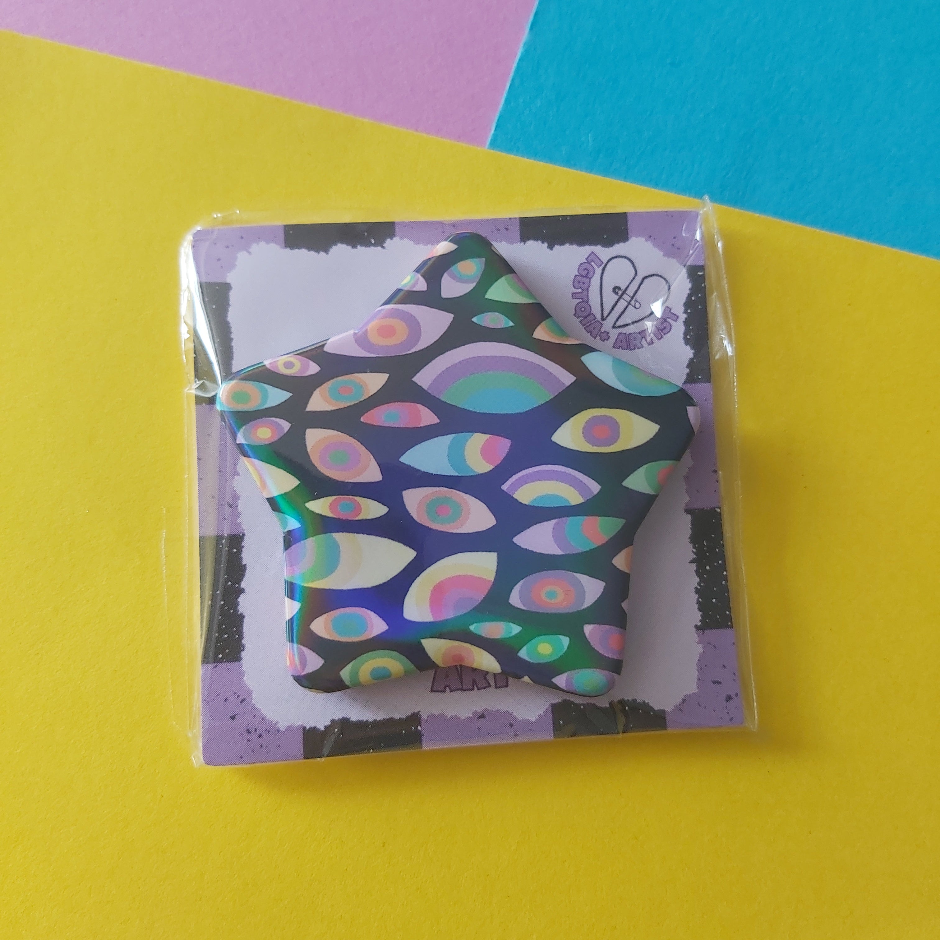 A star-shaped pin with a psychedelic multicolor eye pattern, packaged on a branded backing card and placed on a bright yellow surface with pink and blue paper accents.