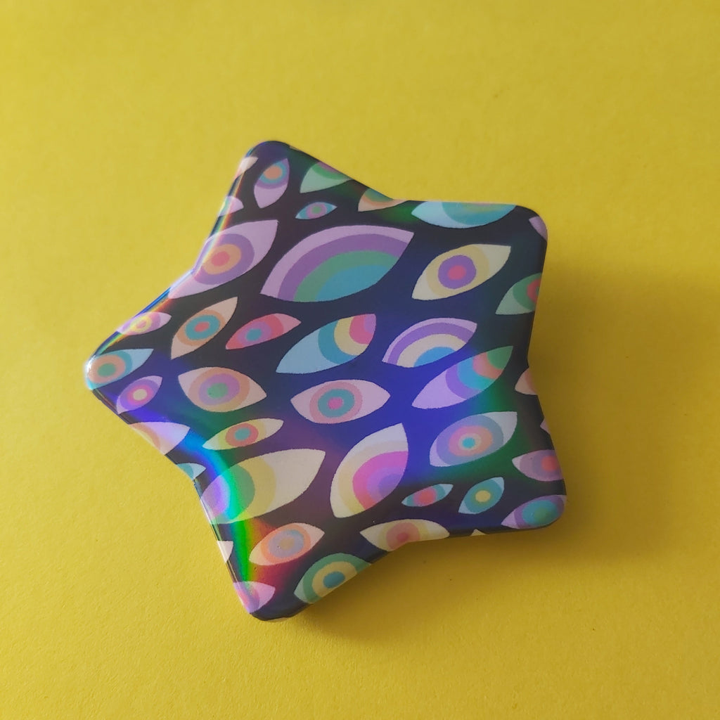A star-shaped pin badge with a colourful, iridescent eye pattern on a dark background, resting on a bright yellow surface.